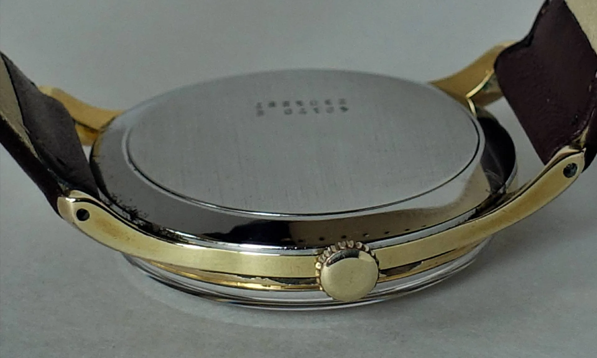 Uhrboden silberfarben Rückseite einer silberfarbenen Armbanduhr mit goldfarbener Krone und braunem Armband.