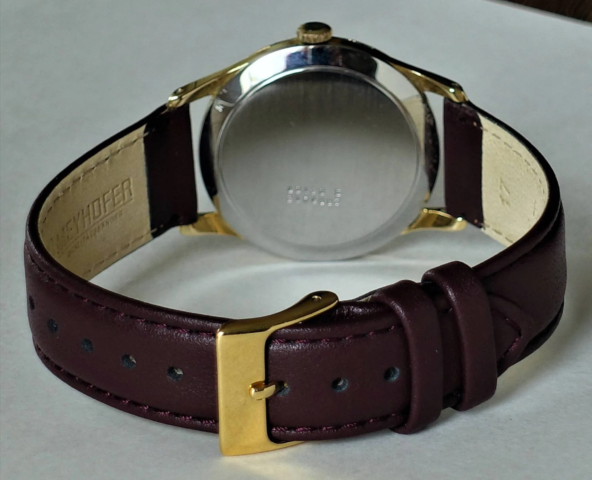 Armbanduhr mit dunkelrotem Lederarmband und goldfarbener Schließe, Rückseite des silberfarbenen Gehäuses sichtbar.