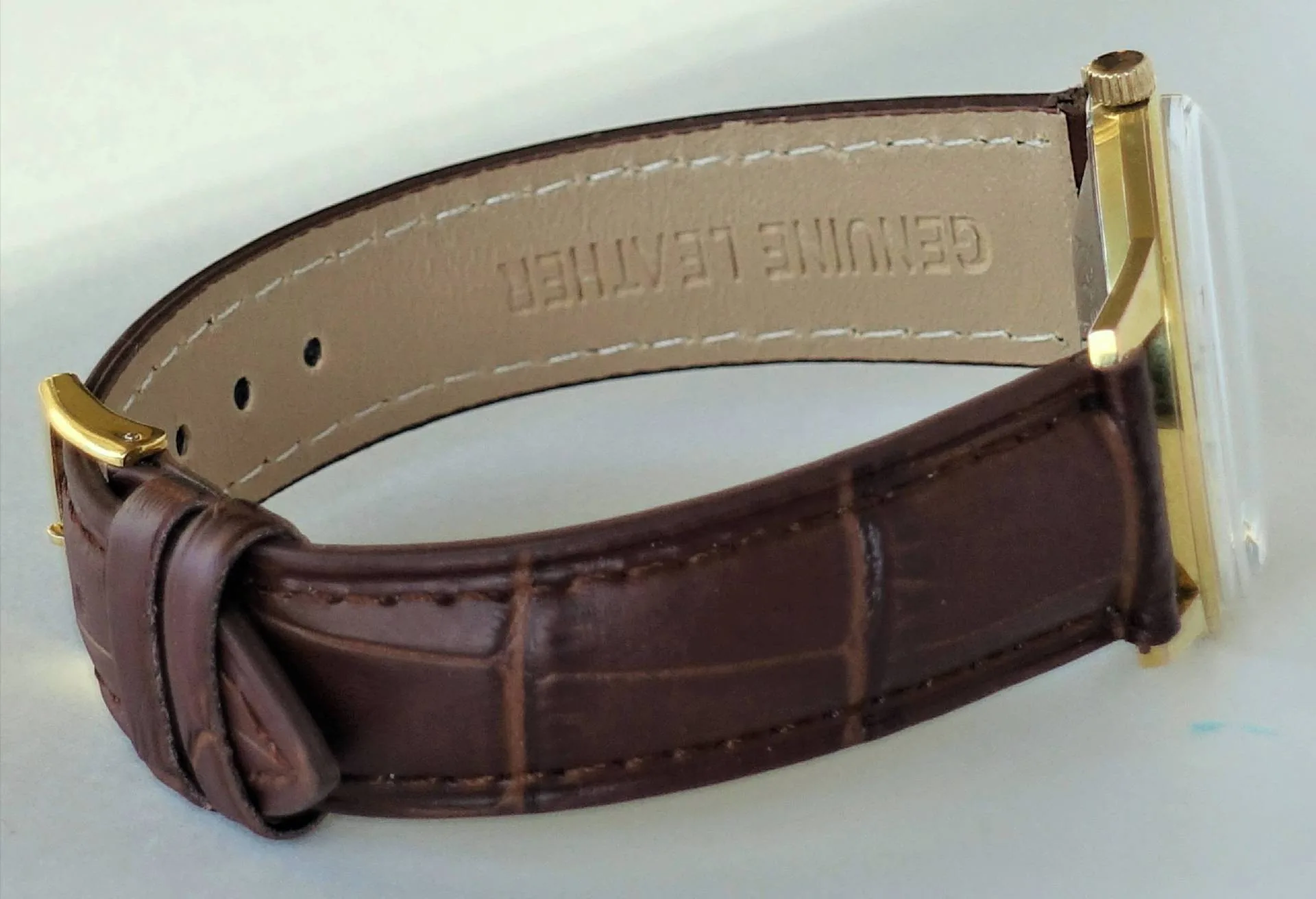 Eine Armbanduhr mit braunem Lederarmband und einem rechteckigen, gelbfarbenen Gehäuse liegt seitlich auf einer weißen Oberfläche.