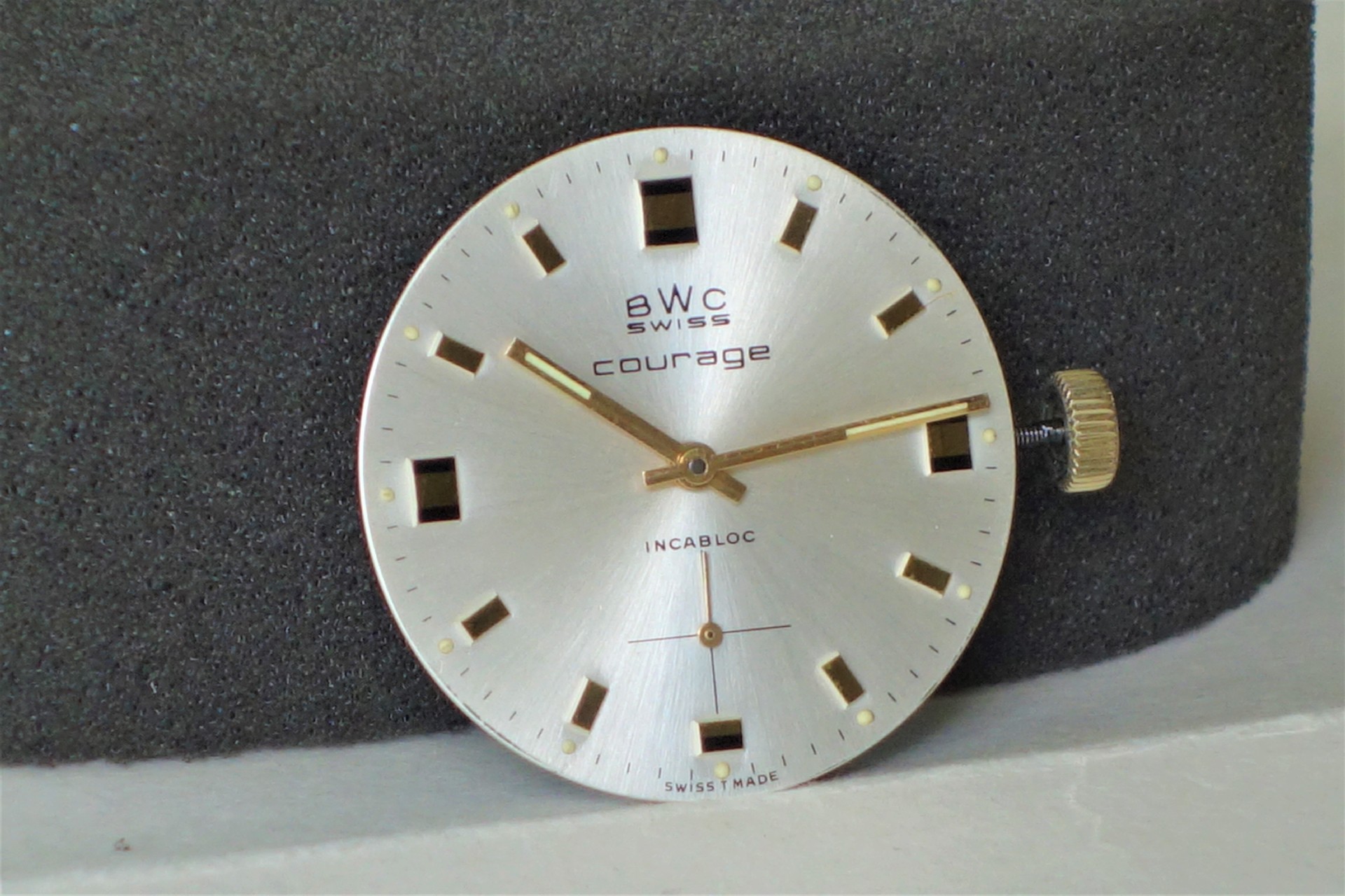 Rundes Uhrwerk mit silberfarbenem Zifferblatt, goldfarbenen Zeigern und Krone, Aufschrift "BWC Swiss courage".