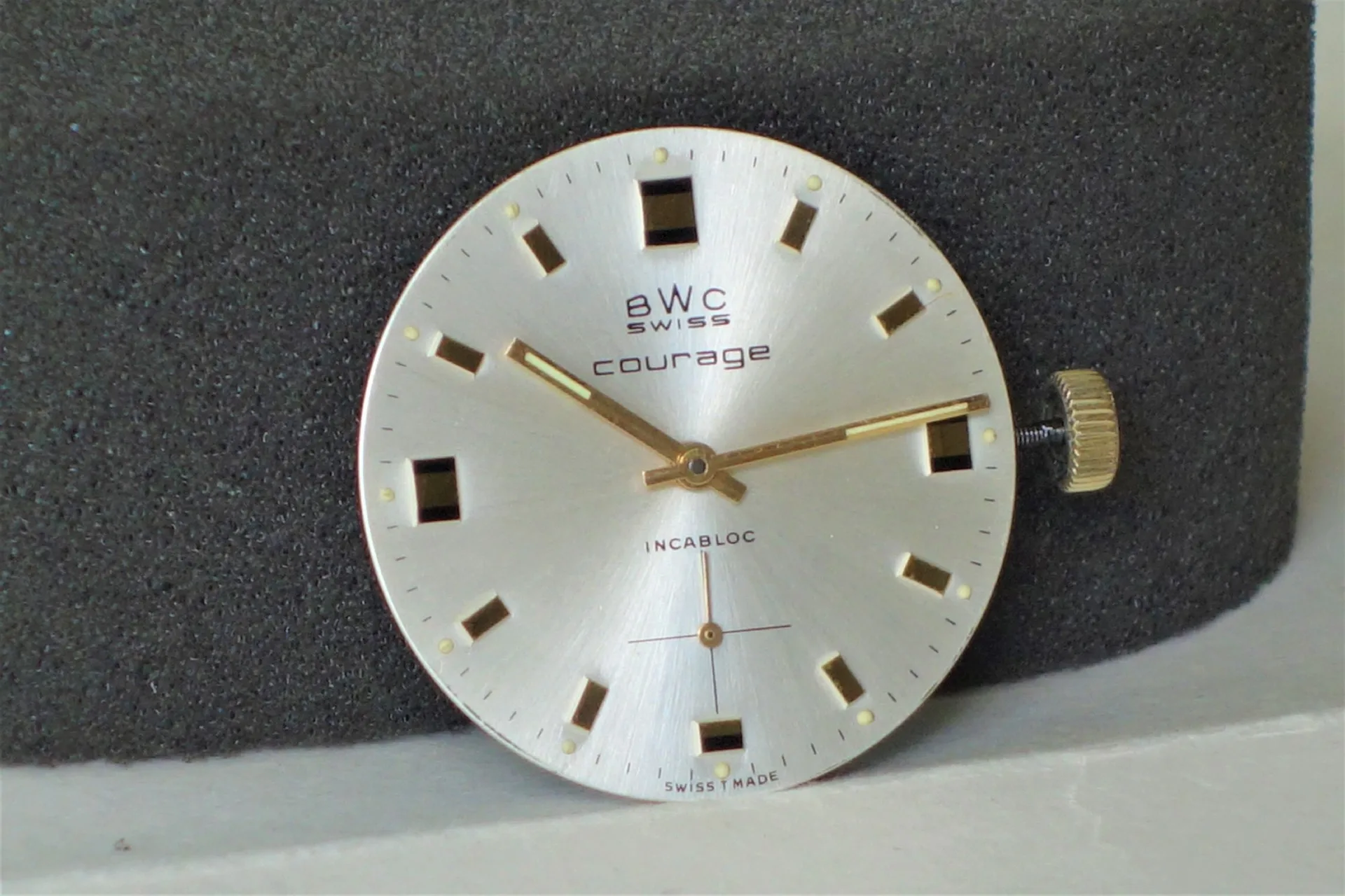 Uhrwerk mit Zifferblatt BWC Rundes Uhrwerk mit silberfarbenem Zifferblatt, goldfarbenen Zeigern und Krone, Aufschrift "BWC Swiss courage".