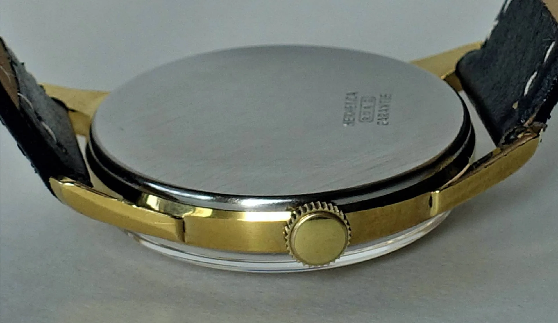 Rundes Armbanduhrgehäuse mit goldfarbener Seite, silberfarbener Rückseite und schwarzem Lederarmband.
