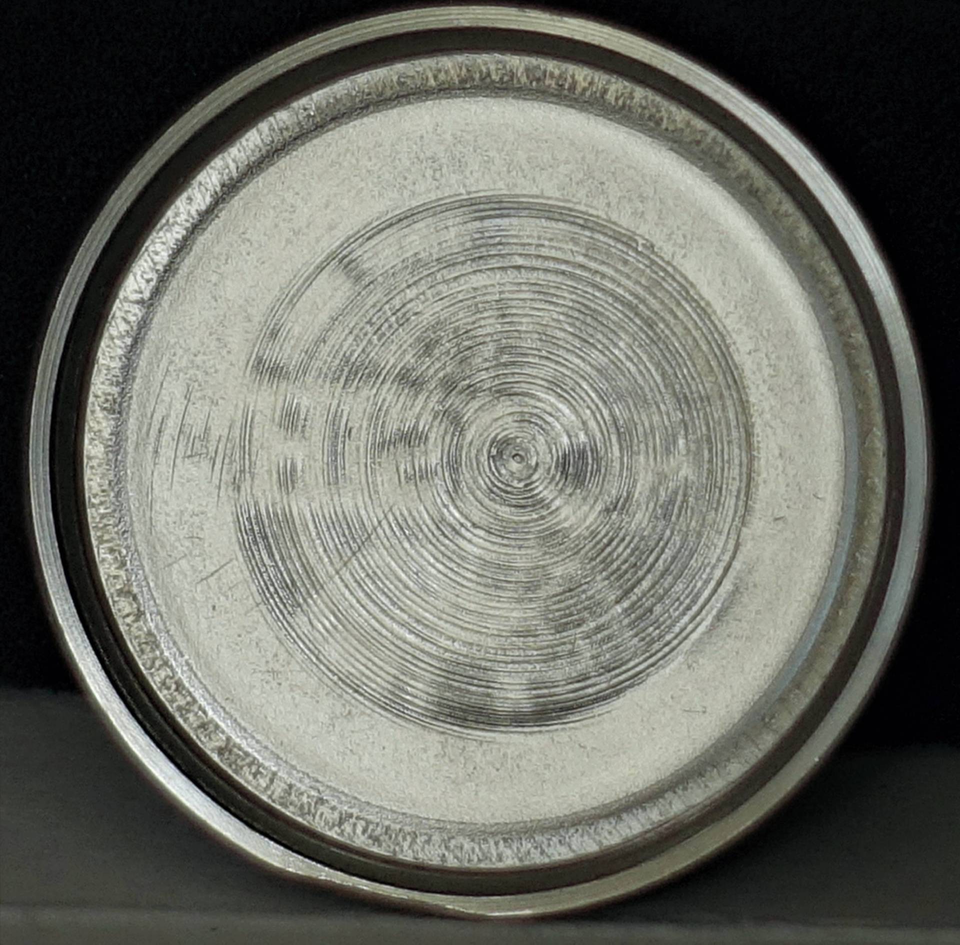 Rundes, silberfarbenes Uhrendeckel-Innenteil mit konzentrischem Schliffmuster, auf schwarzem Hintergrund.