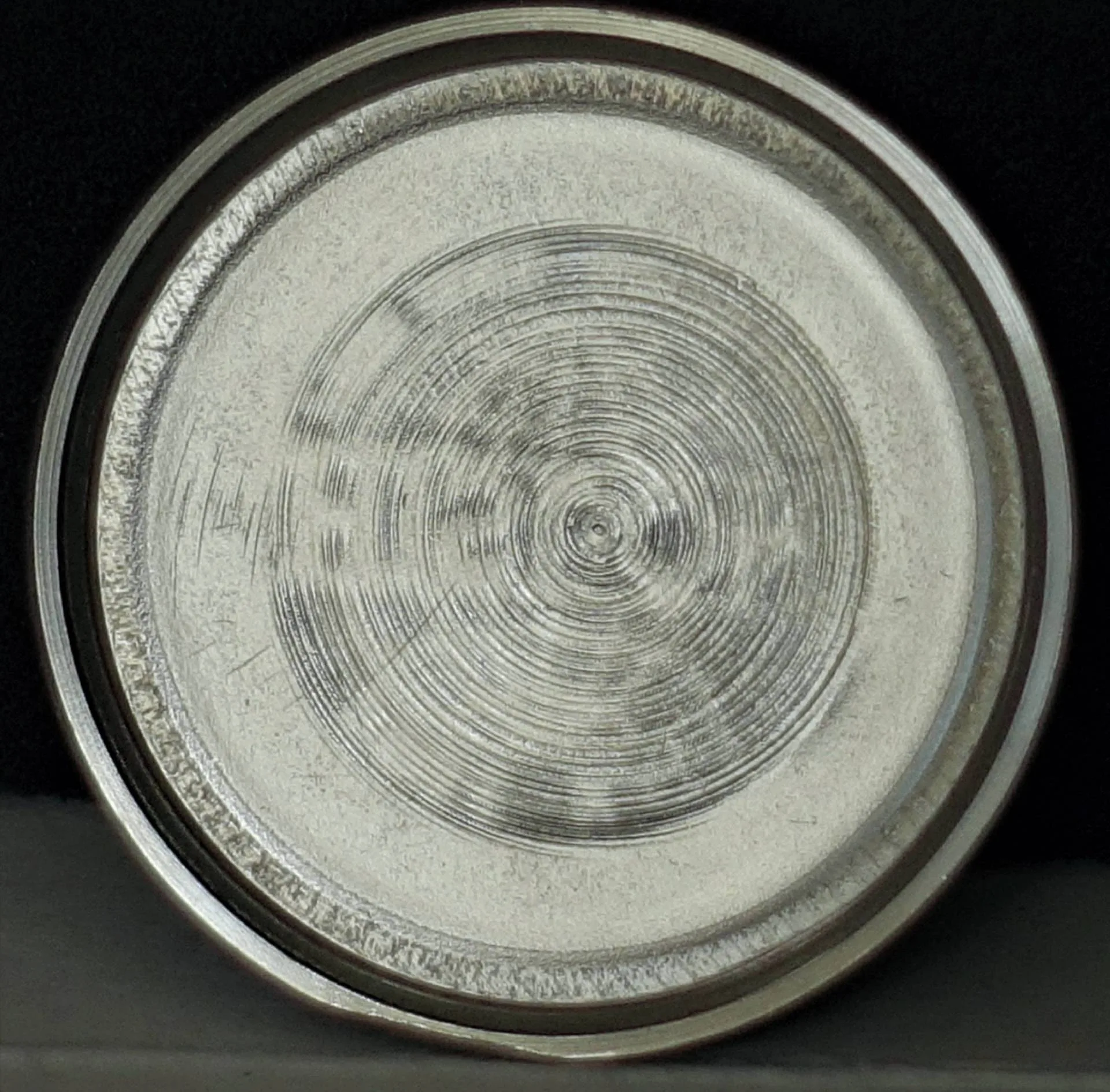 Rundes, silberfarbenes Uhrendeckel-Innenteil mit konzentrischem Schliffmuster, auf schwarzem Hintergrund.