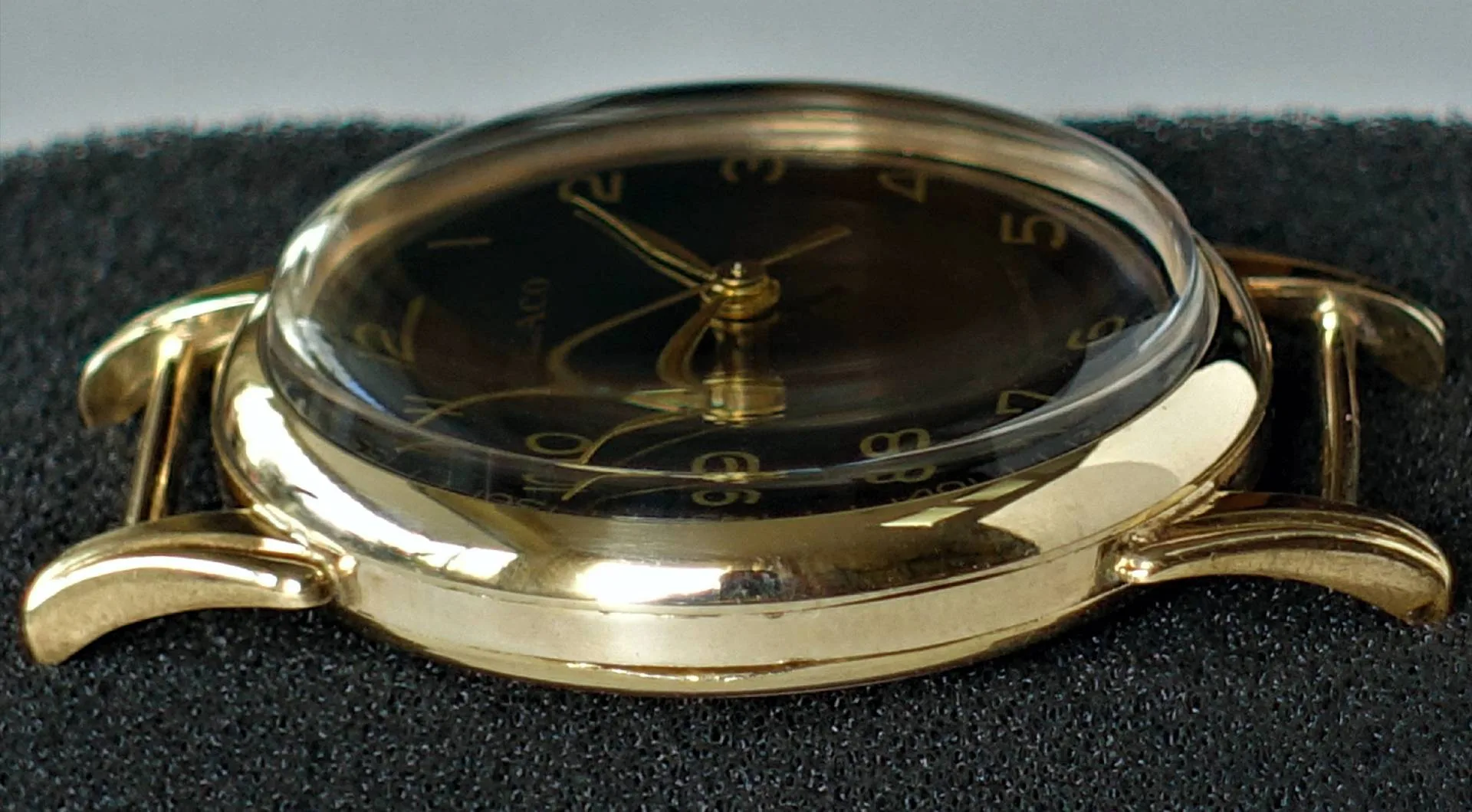 Schwarzes Zifferblatt, gelbes Gehäuse Eine Armbanduhr mit schwarzem Zifferblatt und gelbfarbenem Gehäuse liegt auf schwarzem Schaumstoff, das Glas ist stark gewölbt.