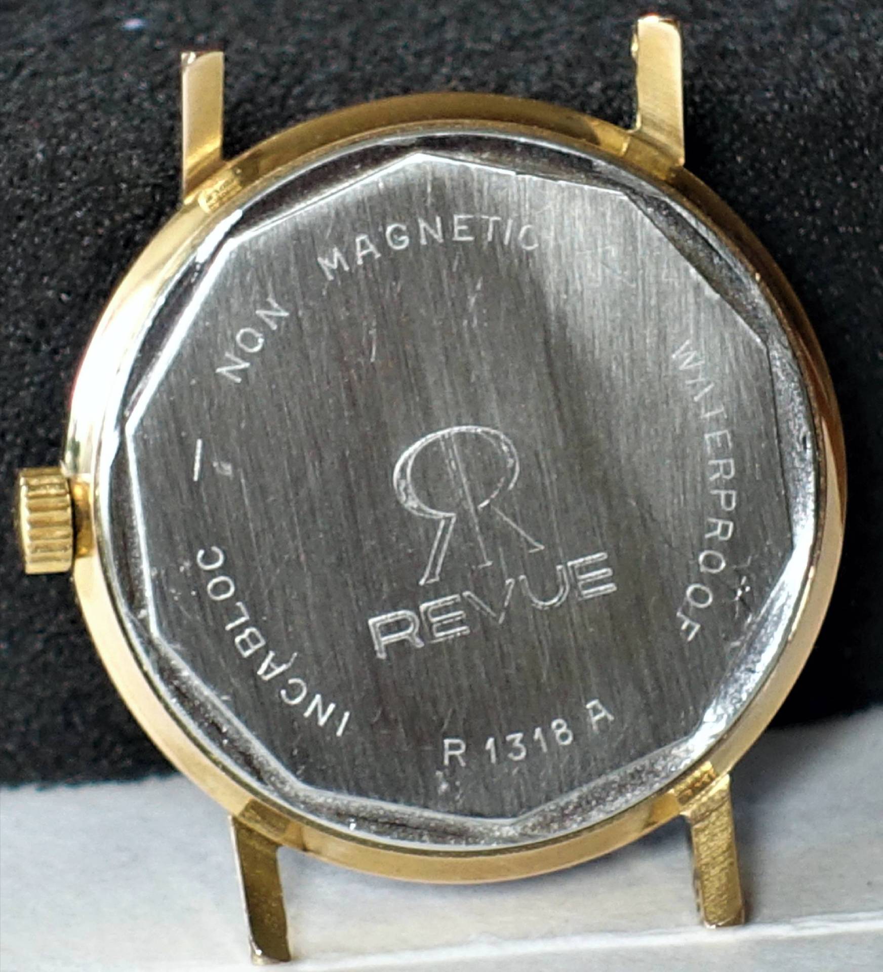Zweifarbiges Uhrgehäuse mit silberfarbener Rückseite und goldfarbenem Rand, Gravur "REVUE" auf dem Boden.