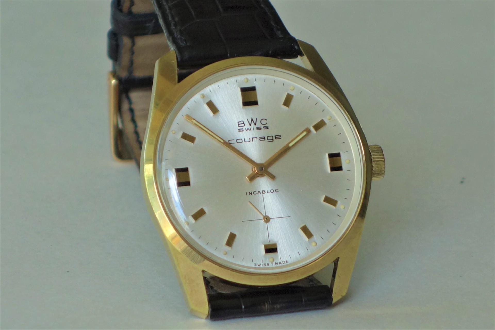 Analoge Armbanduhr mit goldfarbenem Gehäuse, silbernem Zifferblatt und schwarzem Lederarmband.