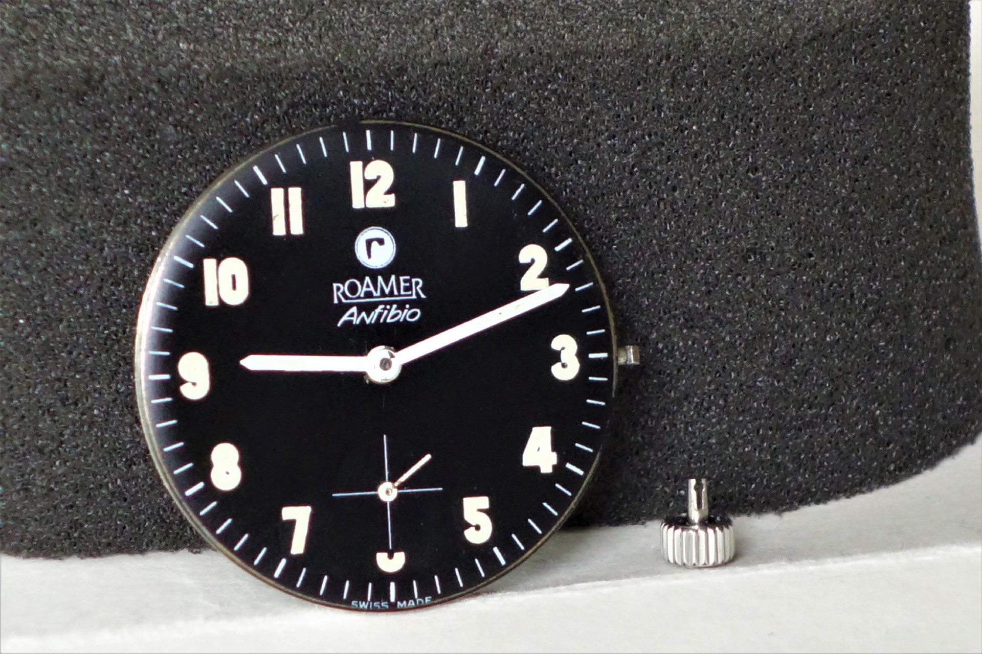 Schwarzes Uhrwerk mit Zifferblatt und Zeigern, daneben eine silberfarbene Krone.