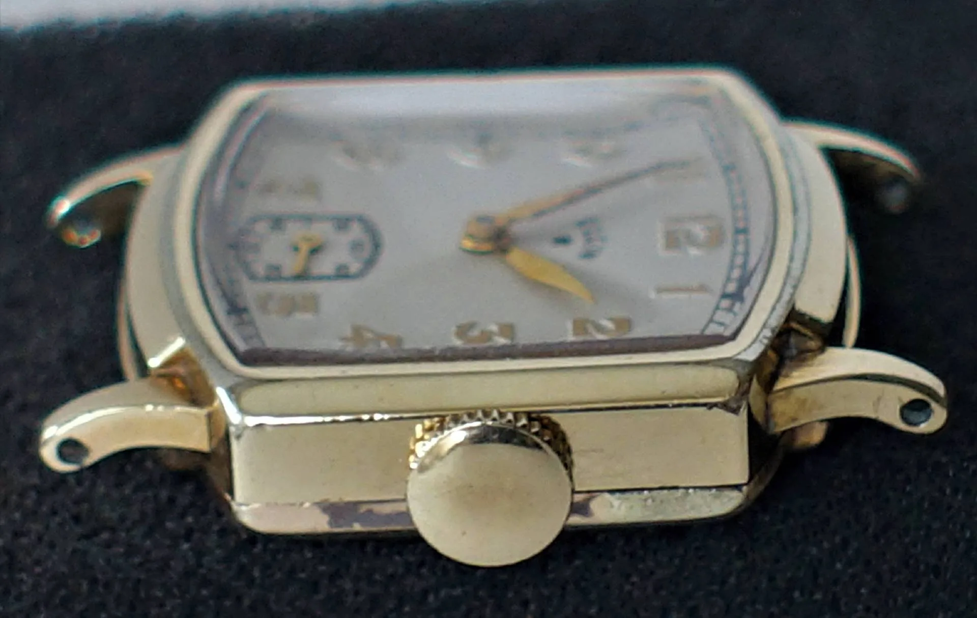 Eine rechteckige Armbanduhr mit gelbgoldenem Gehäuse, goldfarbenen Zeigern und arabischen Ziffern auf einem silberfarbenen Zifferblatt, fotografiert aus der Nähe auf schwarzem Untergrund.