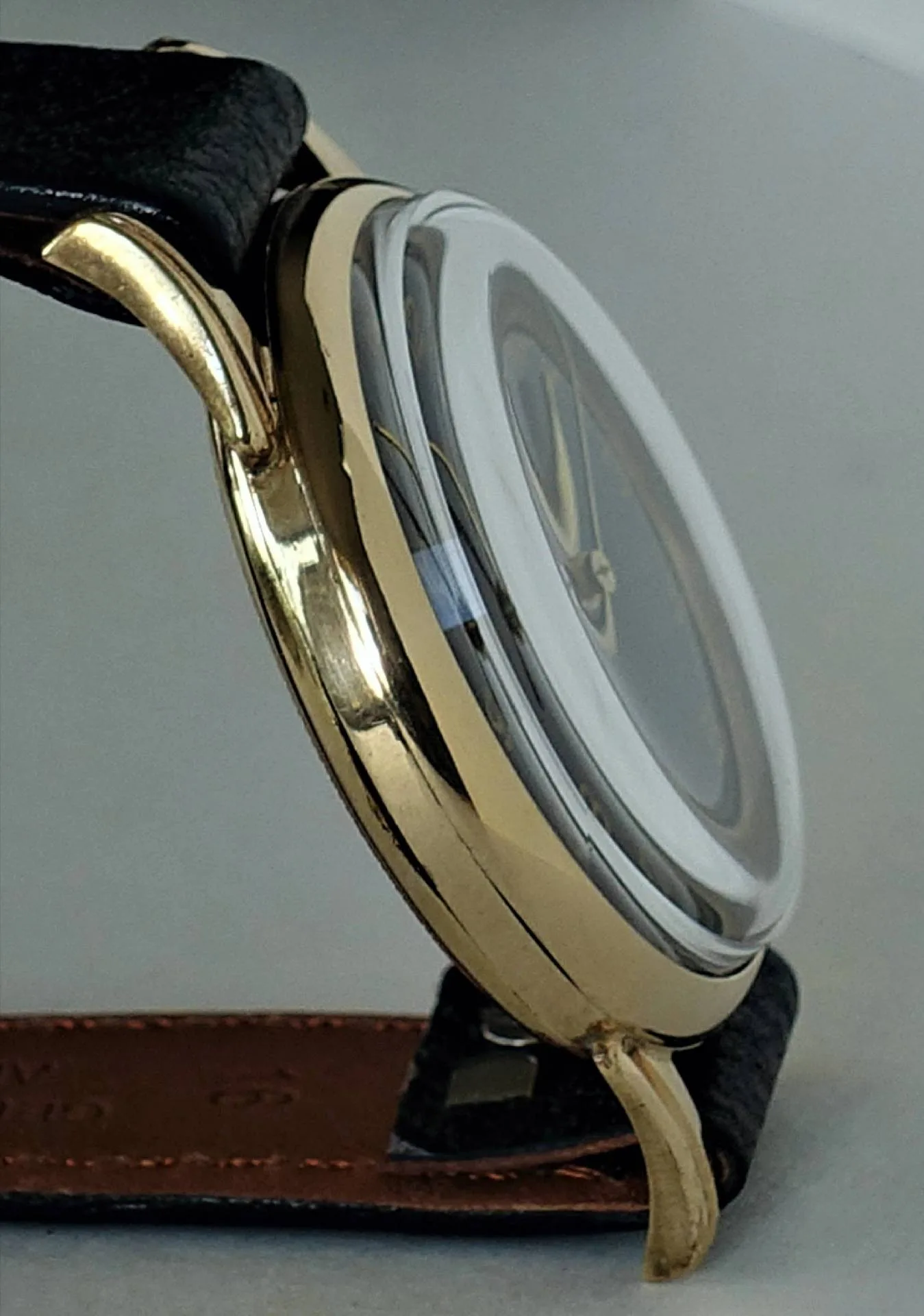 Seitliche Nahaufnahme einer Armbanduhr mit goldfarbenem Gehäuse, schwarzem Band und gewölbtem Glas.