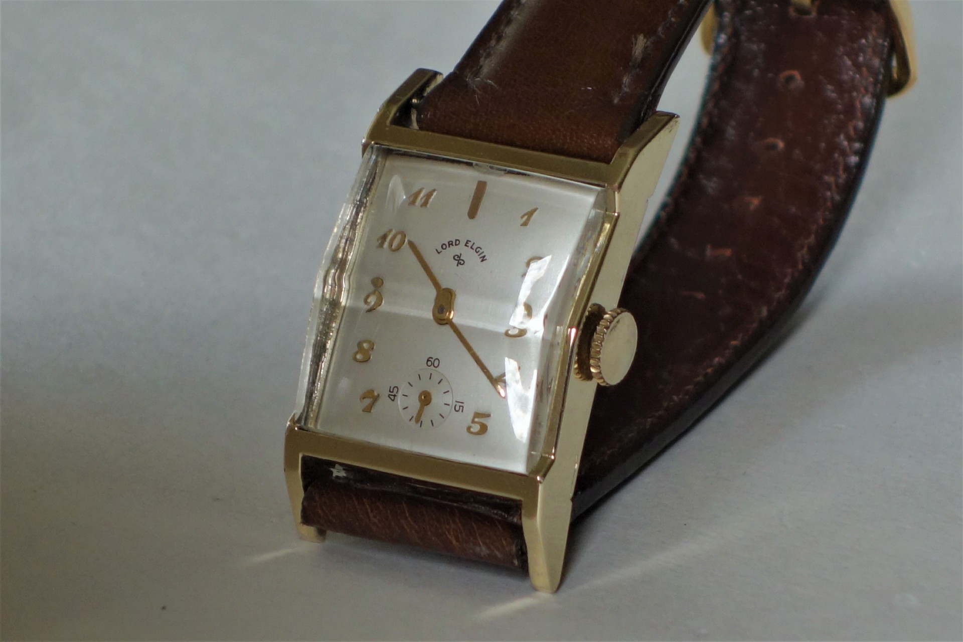 Rechteckige Armbanduhr mit braunem Armband und hellem Zifferblatt.