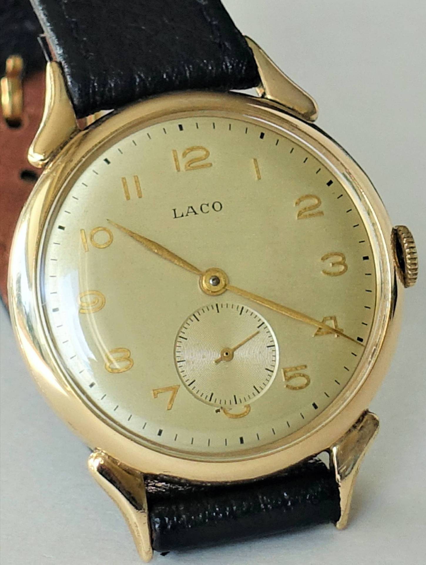 Goldfarbene Armbanduhr mit hellem Zifferblatt, schwarzen Zahlen und schwarzem Lederarmband.