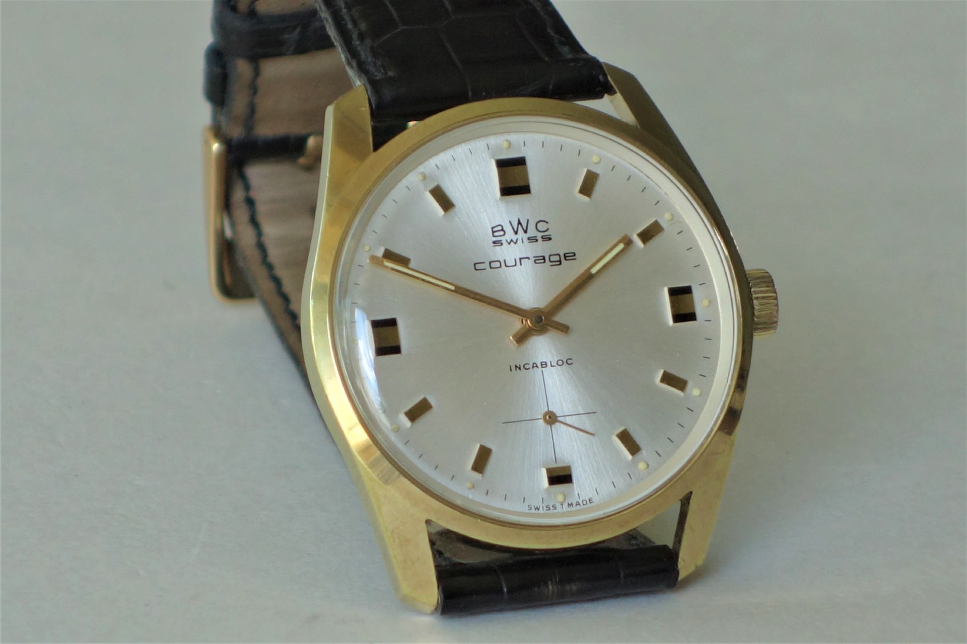 Analoge Armbanduhr mit goldfarbenem Gehäuse, silbernem Zifferblatt und schwarzem Lederarmband.