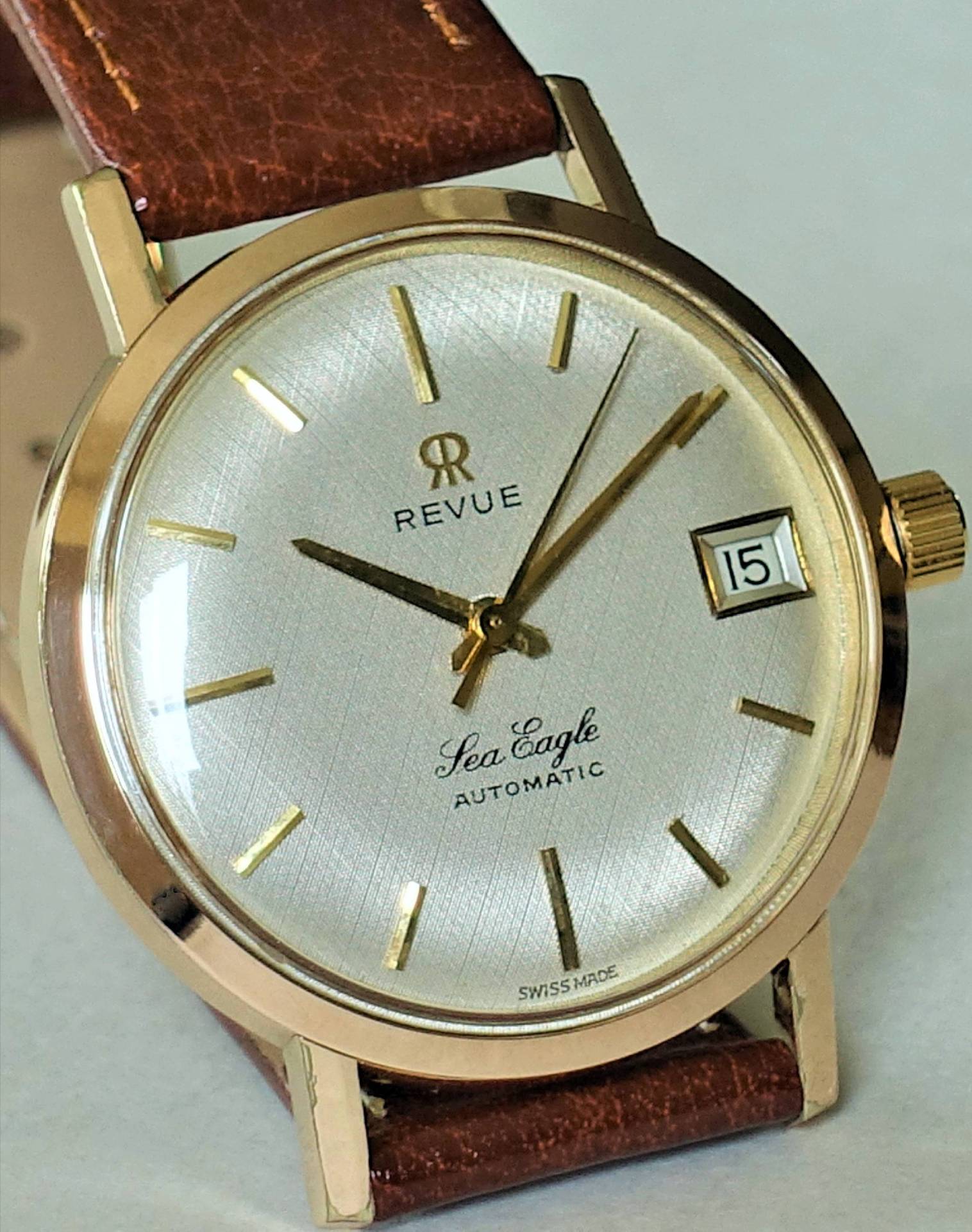 Goldfarbene Armbanduhr mit silbernem Zifferblatt, braunem Armband und Datumsanzeige.