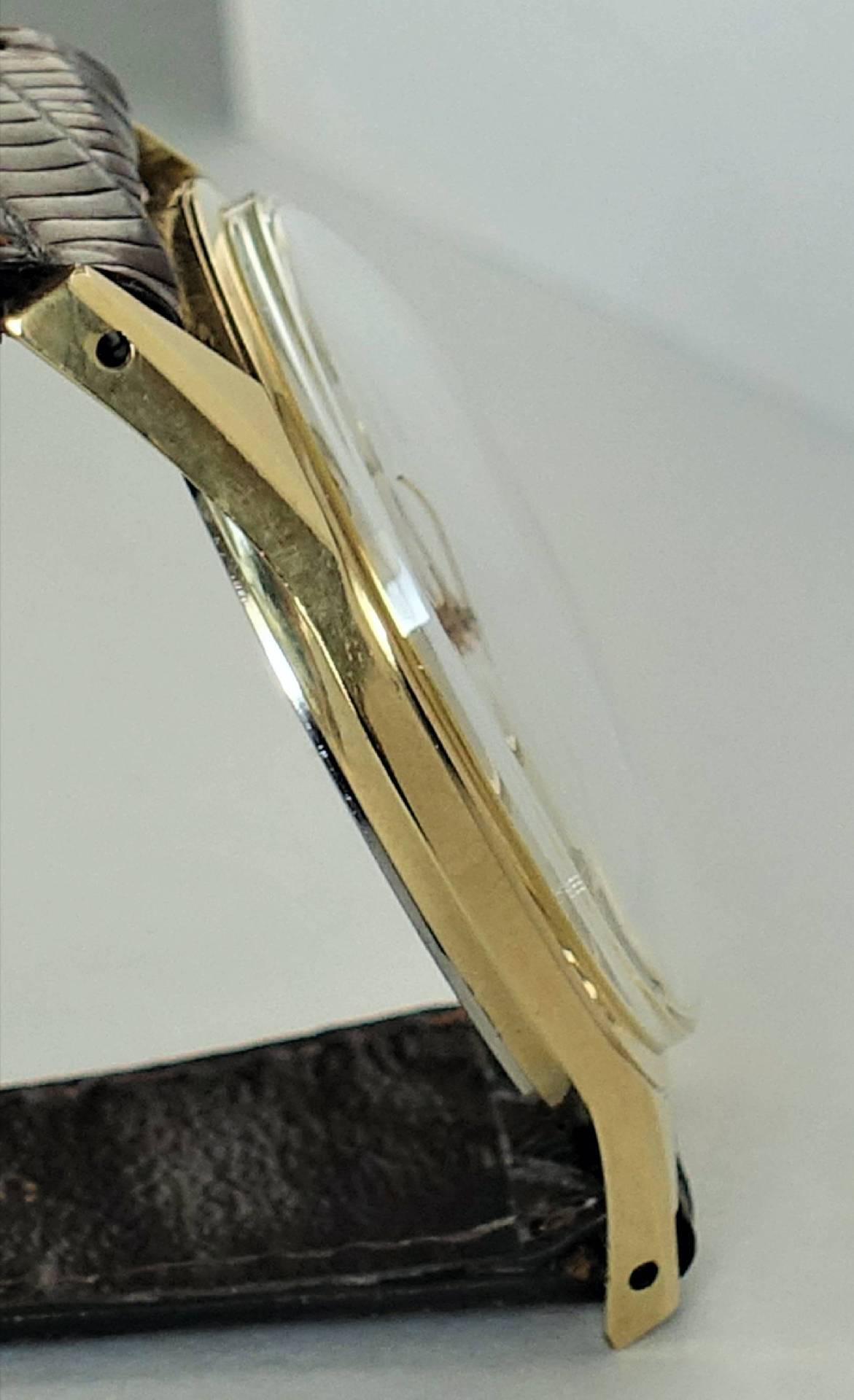 Seitliche Ansicht einer goldfarbenen Armbanduhr mit braunem Armband und gewölbtem Uhrglas.