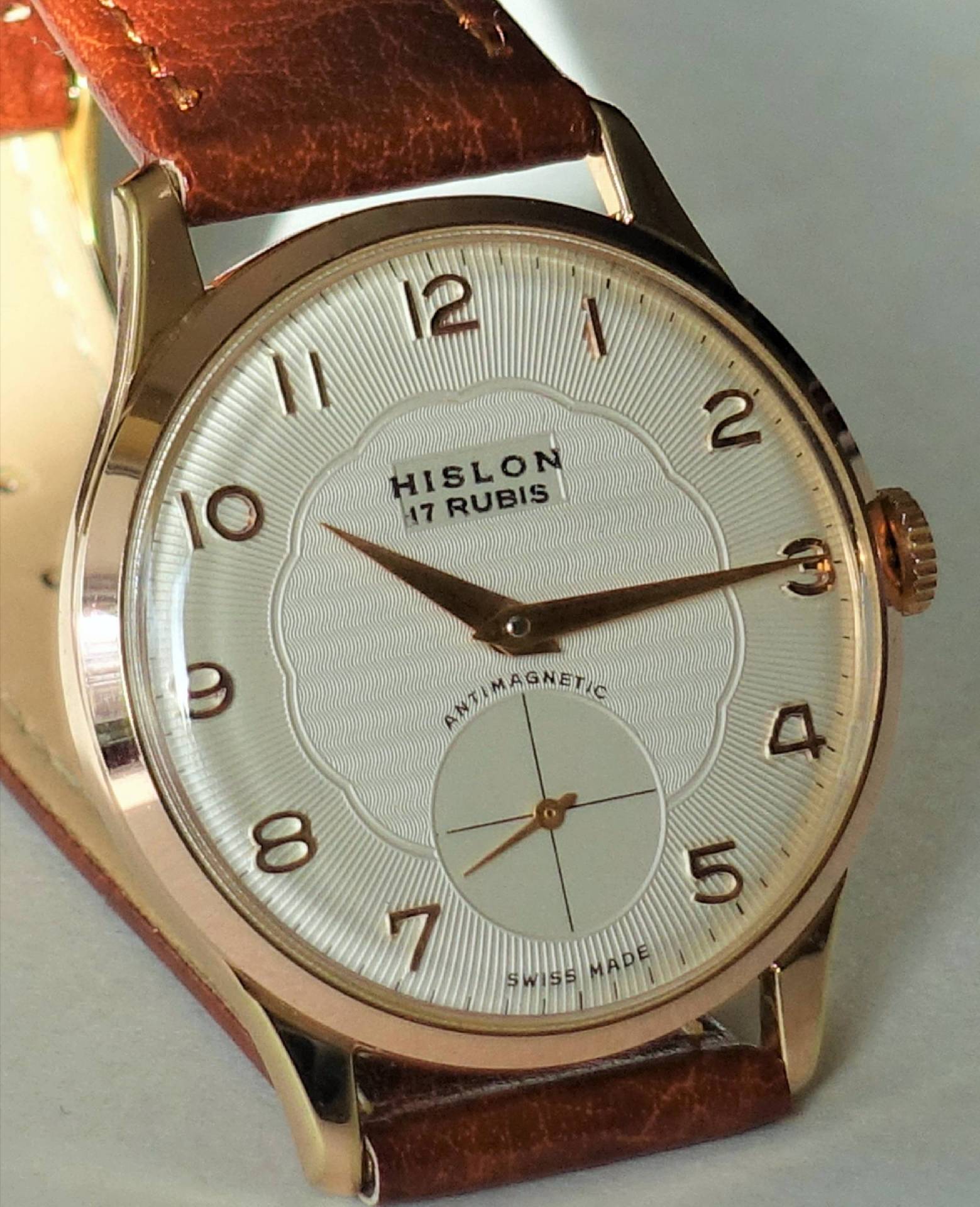 Eine Armbanduhr mit weißem Zifferblatt, bronzefarbenem Gehäuse und braunem Lederarmband, die Marke Hislon und die Aufschrift "17 Rubis" sind auf dem Zifferblatt zu sehen.