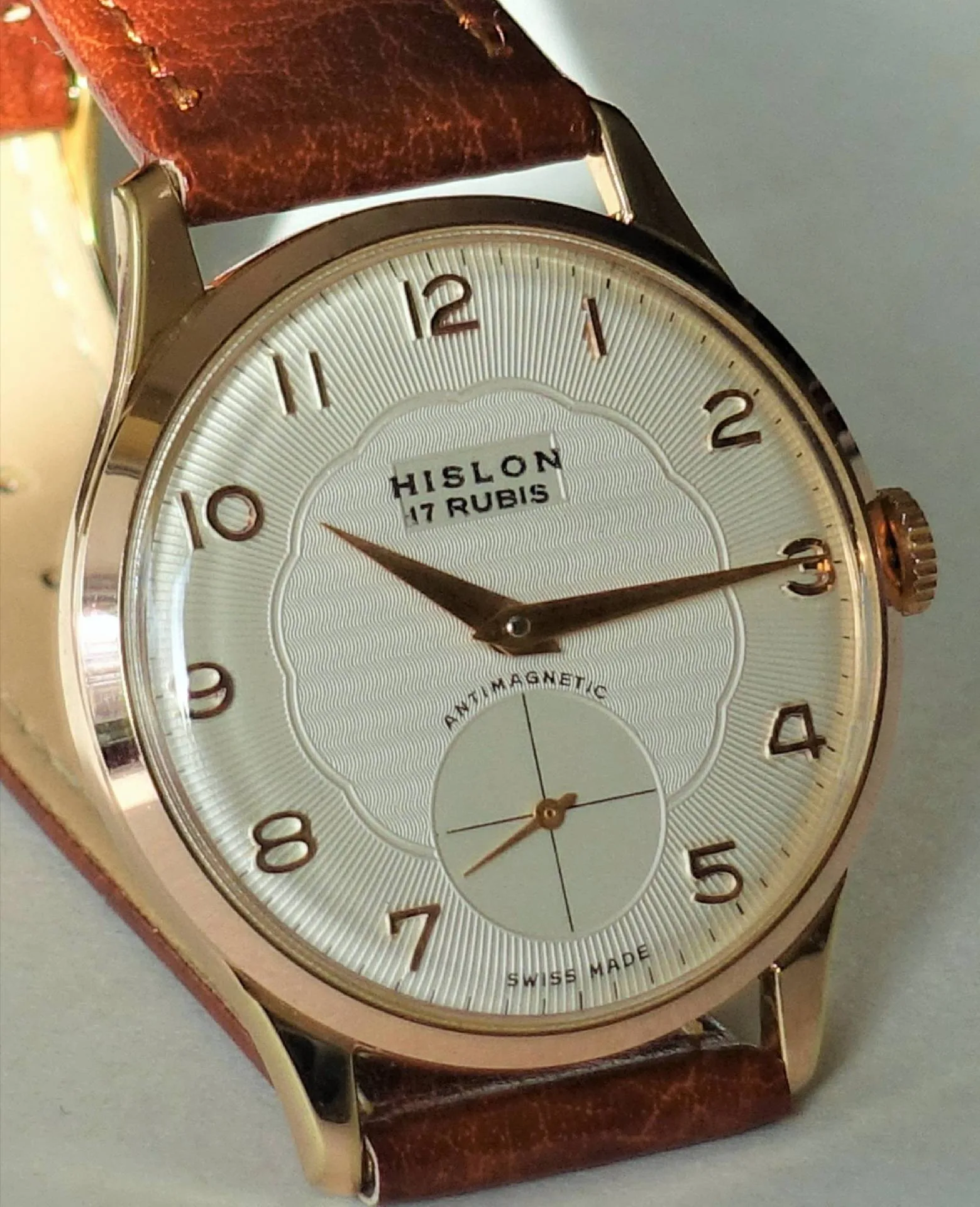 Weißes Zifferblatt, bronzefarbenes Gehäuse Eine Armbanduhr mit weißem Zifferblatt, bronzefarbenem Gehäuse und braunem Lederarmband, die Marke Hislon und die Aufschrift "17 Rubis" sind auf dem Zifferblatt zu sehen.