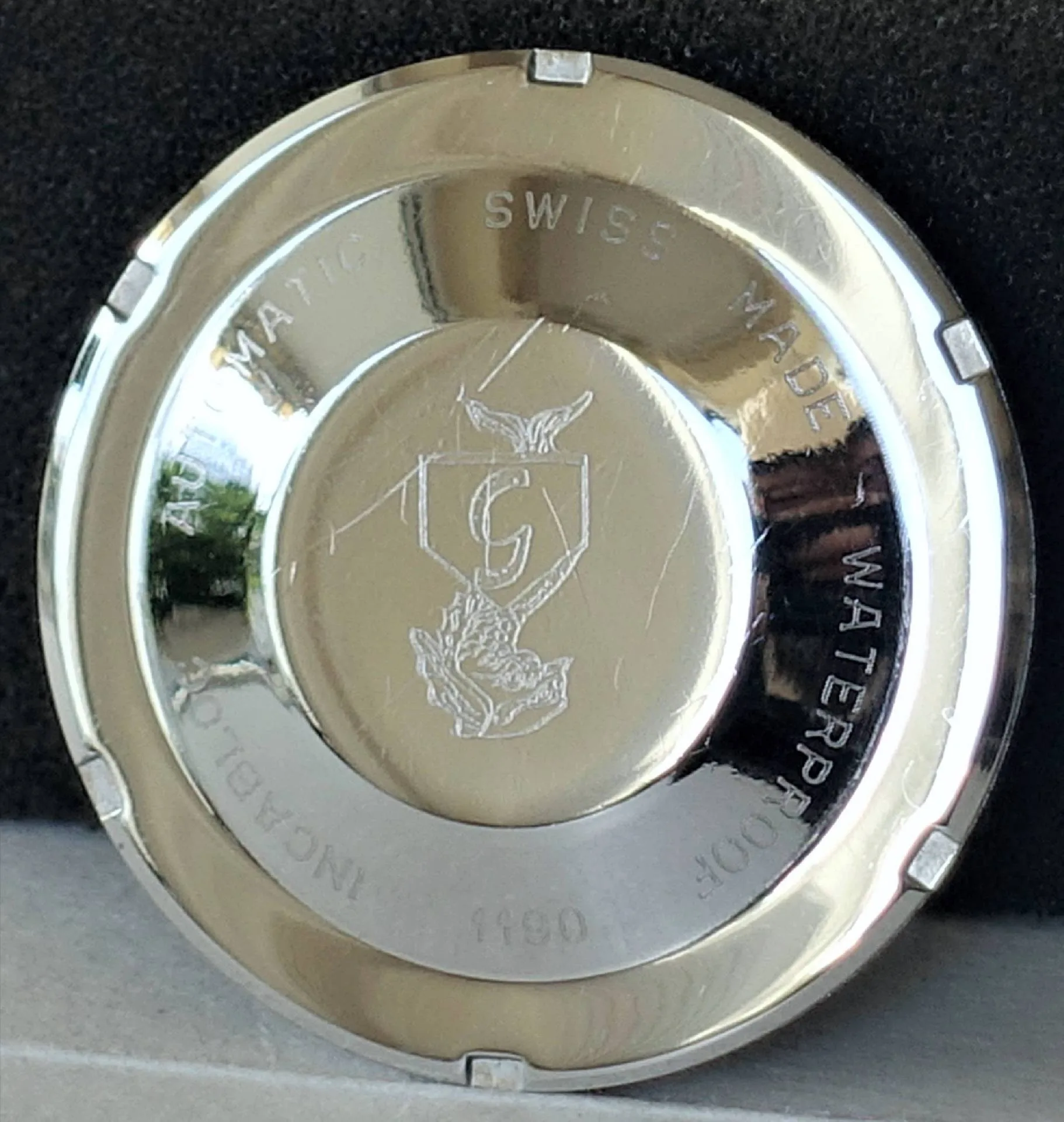 Silberfarbener Uhrenboden mit Gravur Rückseite eines silberfarbenen Uhrenbodens mit Gravur eines Wappens, umlaufender Schrift und polierter Oberfläche.