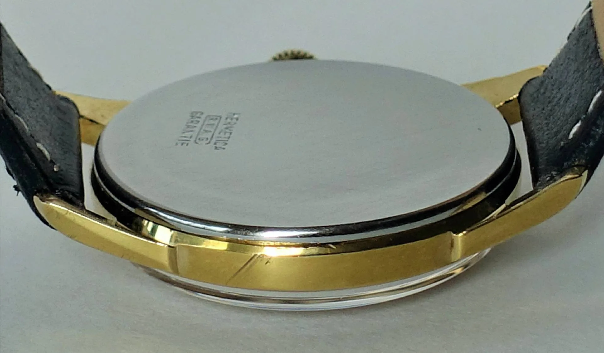 Silberfarbener Uhrboden einer Armbanduhr mit goldfarbenem Gehäuse und schwarzem Armband.