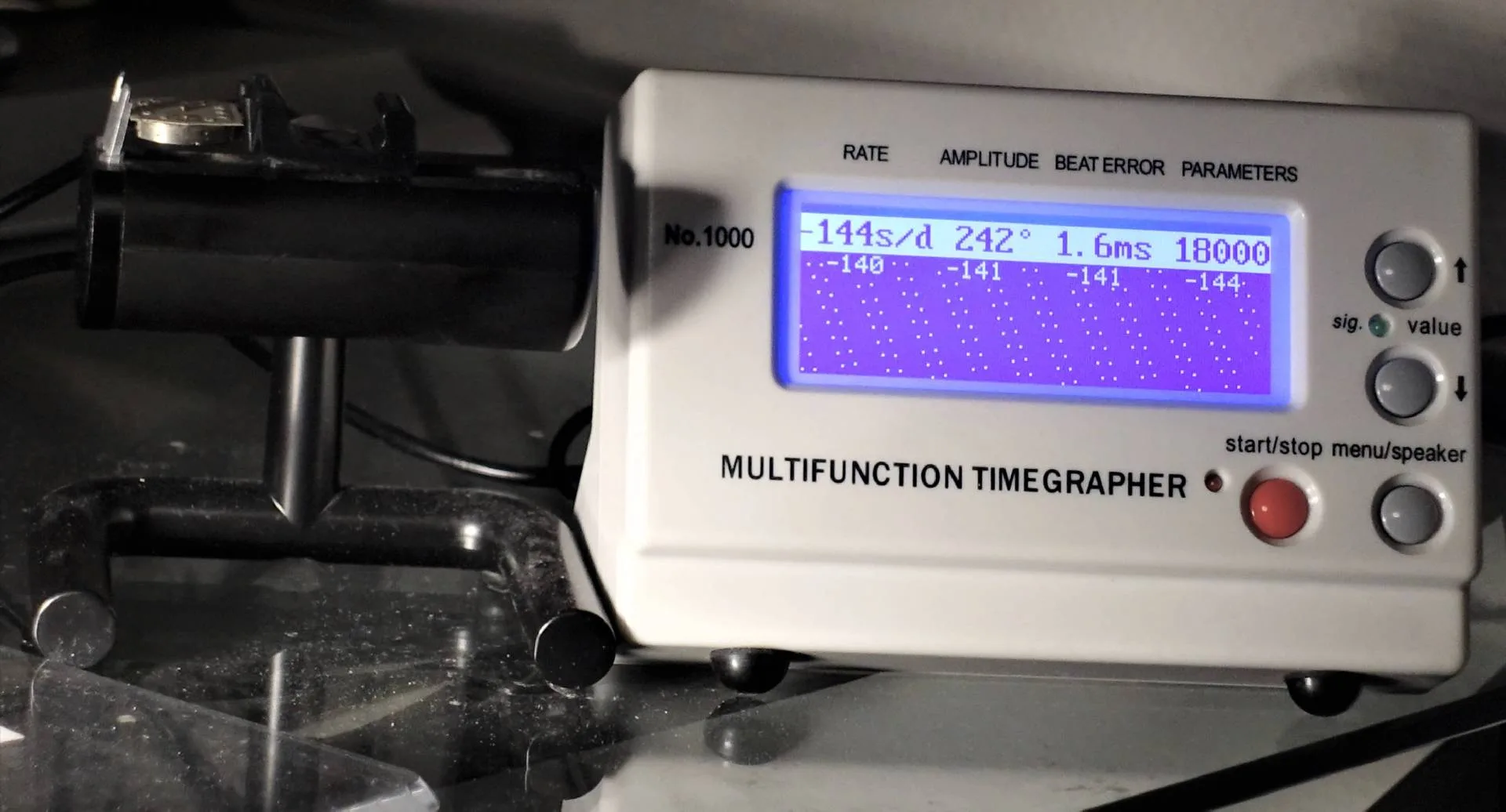 Ein Multifunktions-Zeitmessgerät für mechanische Uhren zeigt Messwerte auf einem leuchtend blauen Display an, daneben liegt eine Uhr in einer schwarzen Halterung.