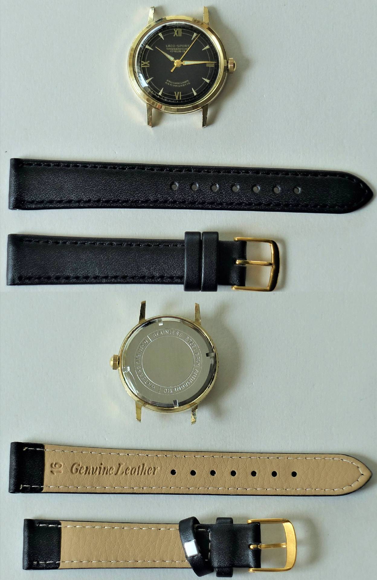 Zerlegte Armbanduhr mit goldfarbenem Gehäuse, schwarzem Zifferblatt und zwei schwarzen Lederarmbändern mit goldfarbener Schließe.