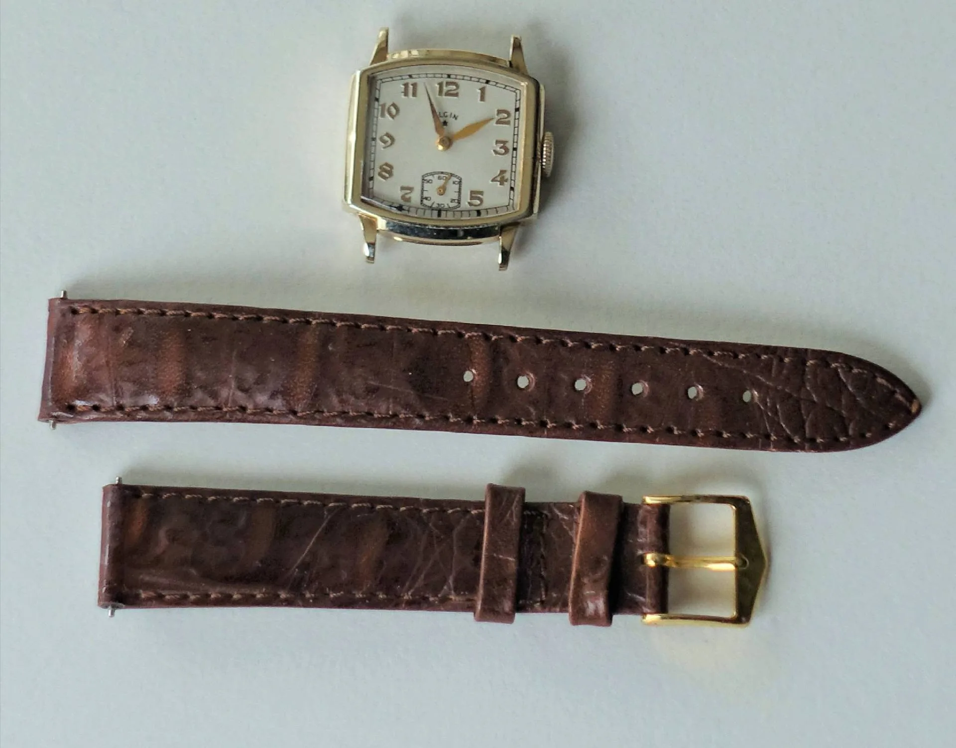 Beige Uhr, braunes Lederband Eine beige Armbanduhr mit eckigem Gehäuse liegt neben einem zweiteiligen braunen Lederarmband mit goldfarbener Schließe.