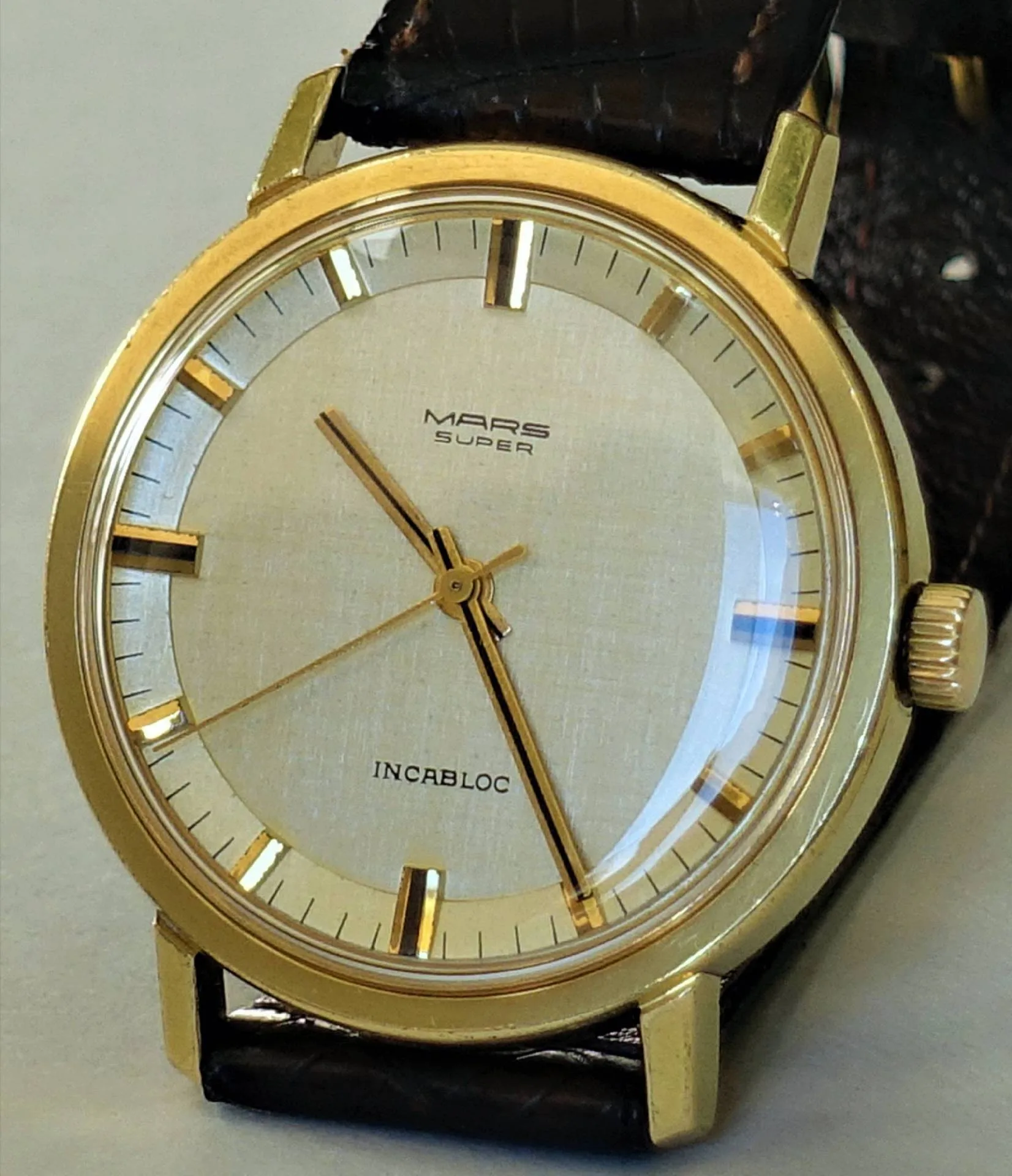 Mars Super Armbanduhr Goldfarbene Armbanduhr mit hellem Zifferblatt und schwarzem Lederarmband.