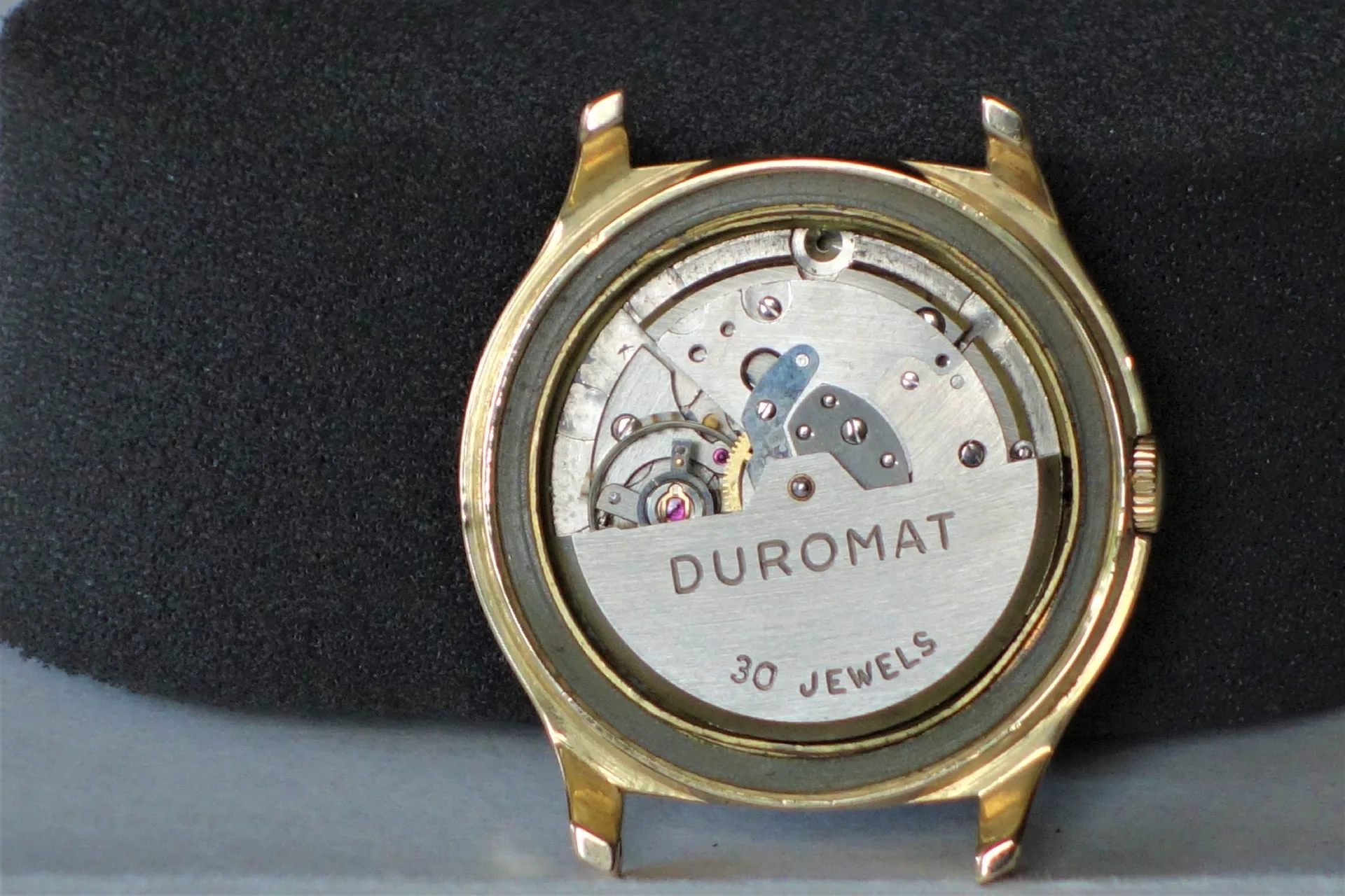 Uhrwerk Duromat 30 Jewels Silberfarbenes mechanisches Uhrwerk mit der Aufschrift "DUROMAT 30 JEWELS" in einem goldfarbenen Uhrengehäuse.