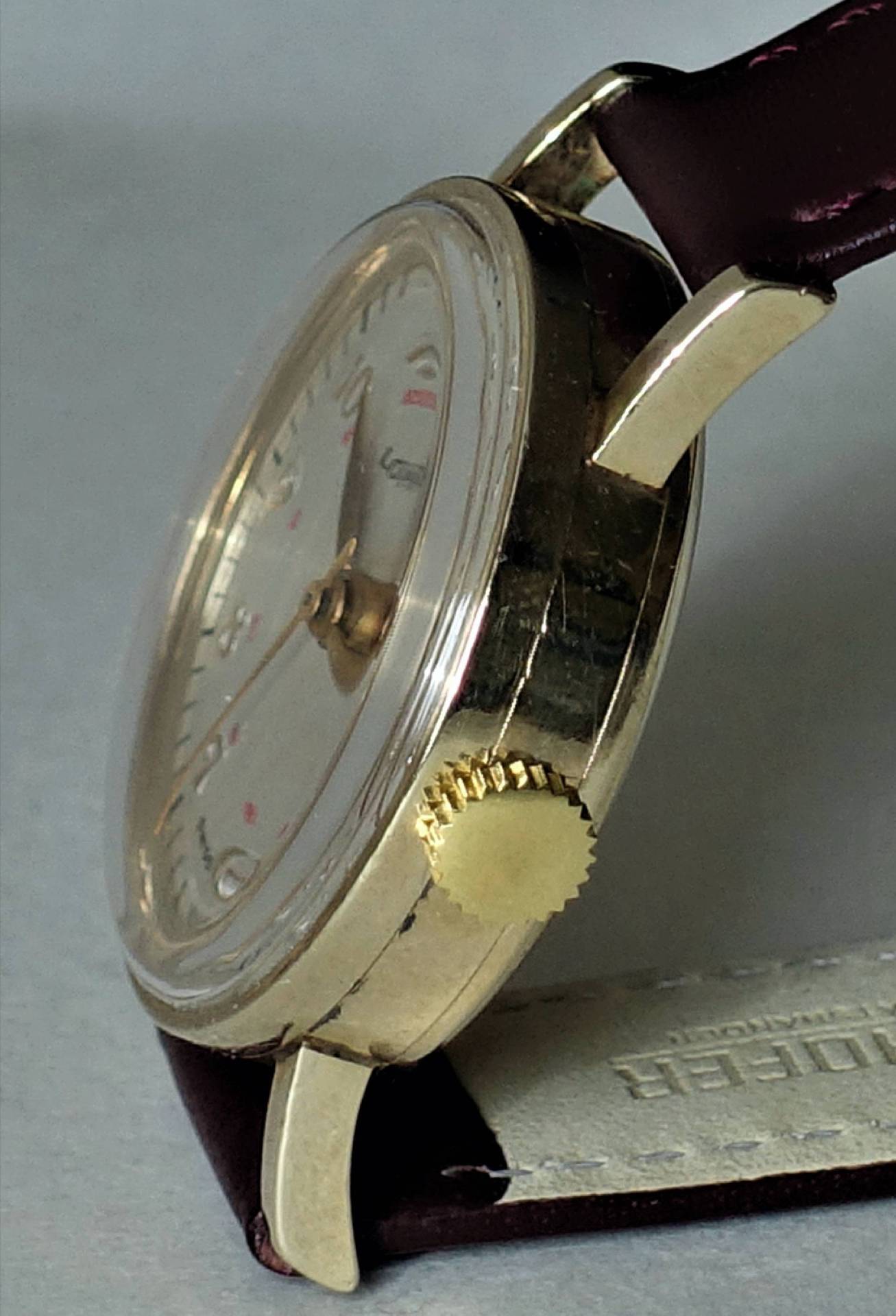 Detailansicht einer goldenen Armbanduhr mit burgunderfarbenem Lederarmband und einem klaren Zifferblatt