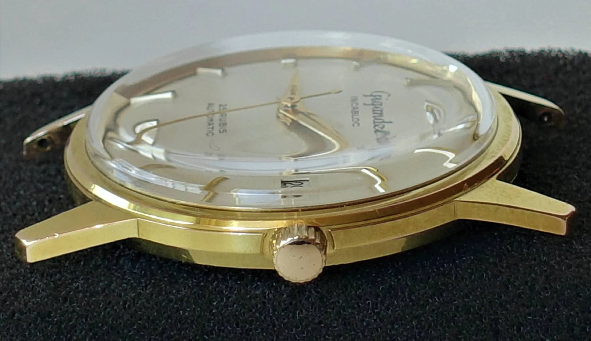 Eine Armbanduhr mit glänzend gelbfarbenem Gehäuse, gewölbtem Glas und hellem Zifferblatt liegt seitlich auf schwarzem Stoff.