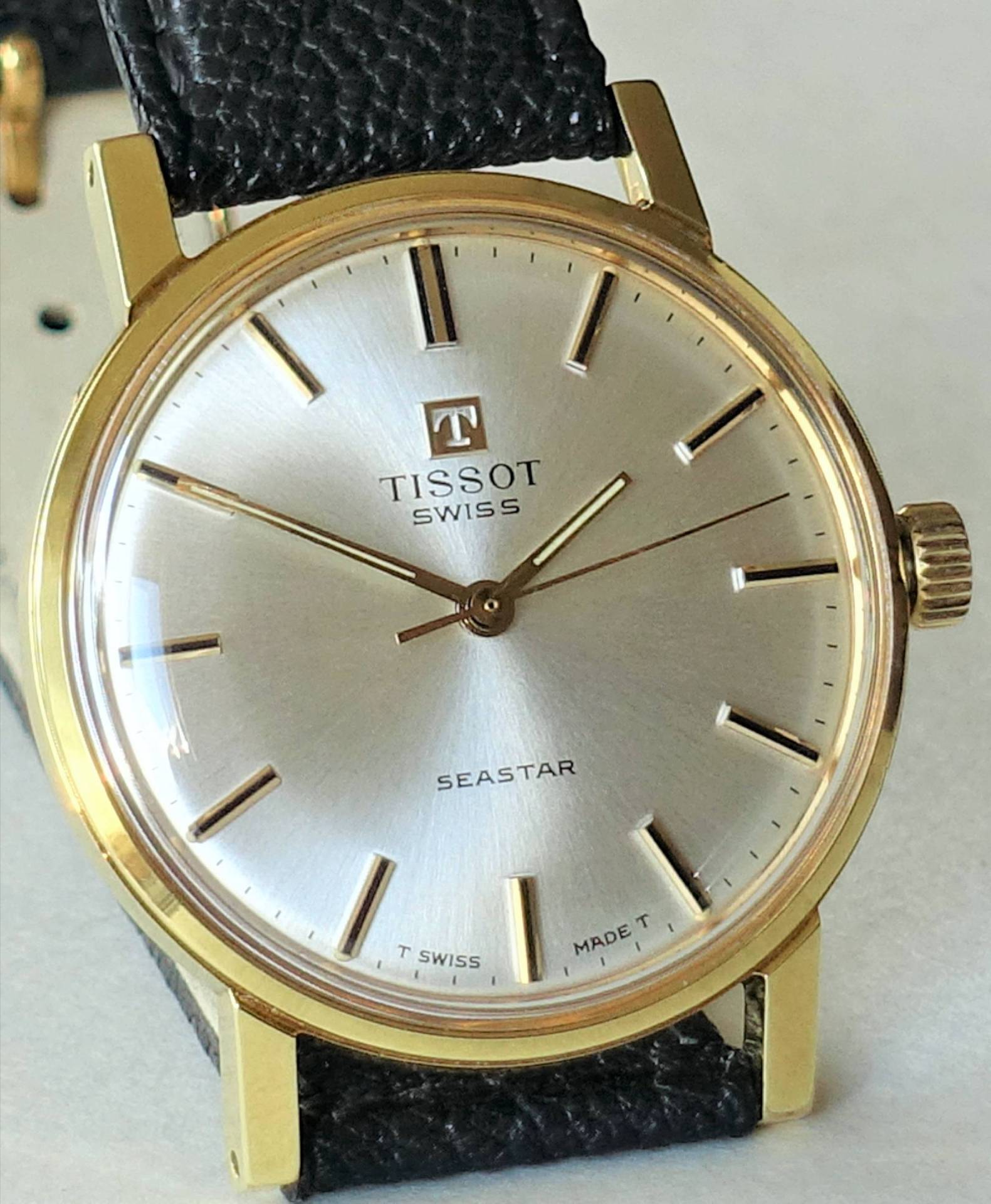 Goldfarbene Armbanduhr mit silberfarbenem Zifferblatt und schwarzem Armband.