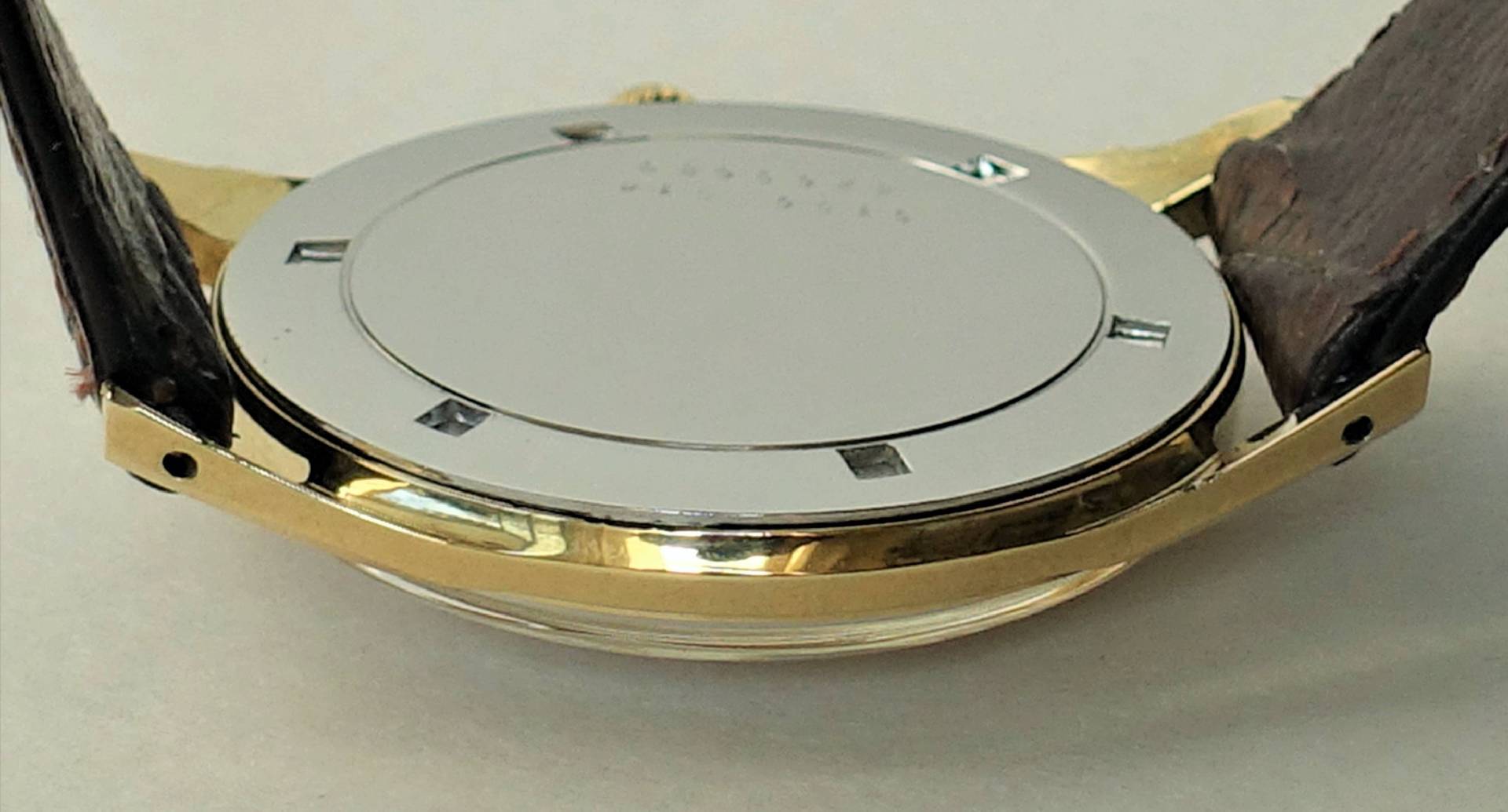 Silberfarbener Uhrenboden an einem Gehäuse mit goldfarbener Umrandung und braunem Armband.