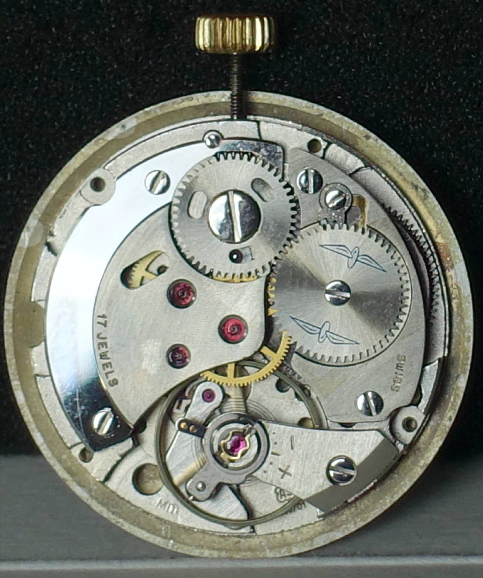 Silberfarbenes mechanisches Uhrwerk mit sichtbaren Zahnrädern und goldfarbener Krone.