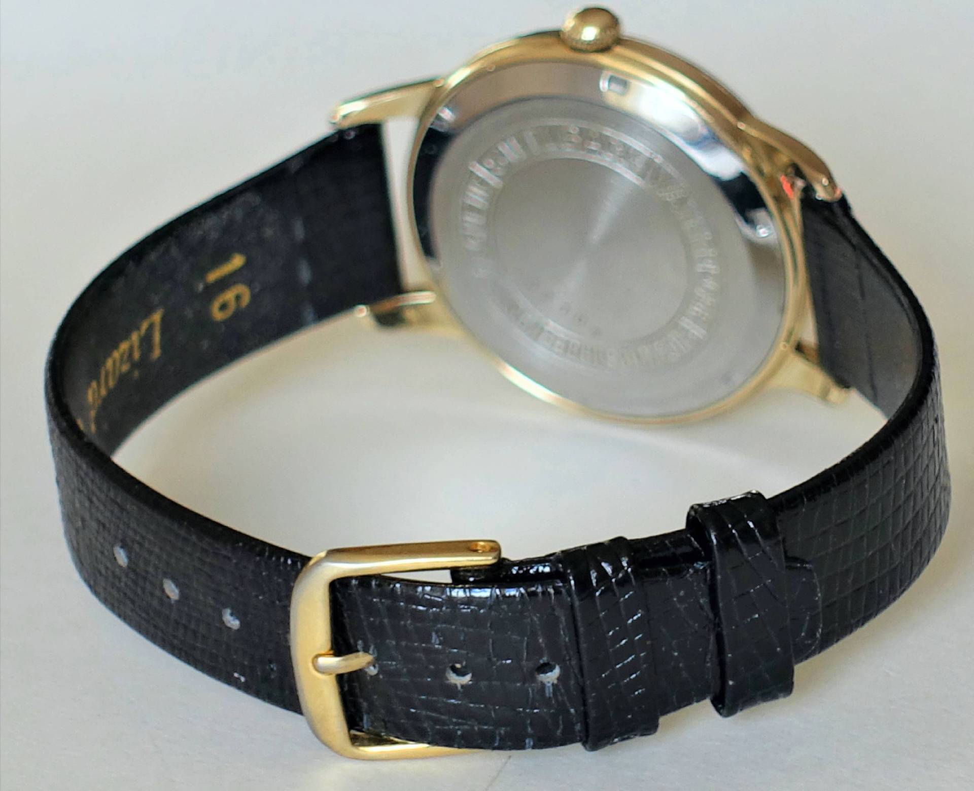 Armbanduhr mit schwarzem Armband und goldfarbenem Gehäuse, Rückseite sichtbar.