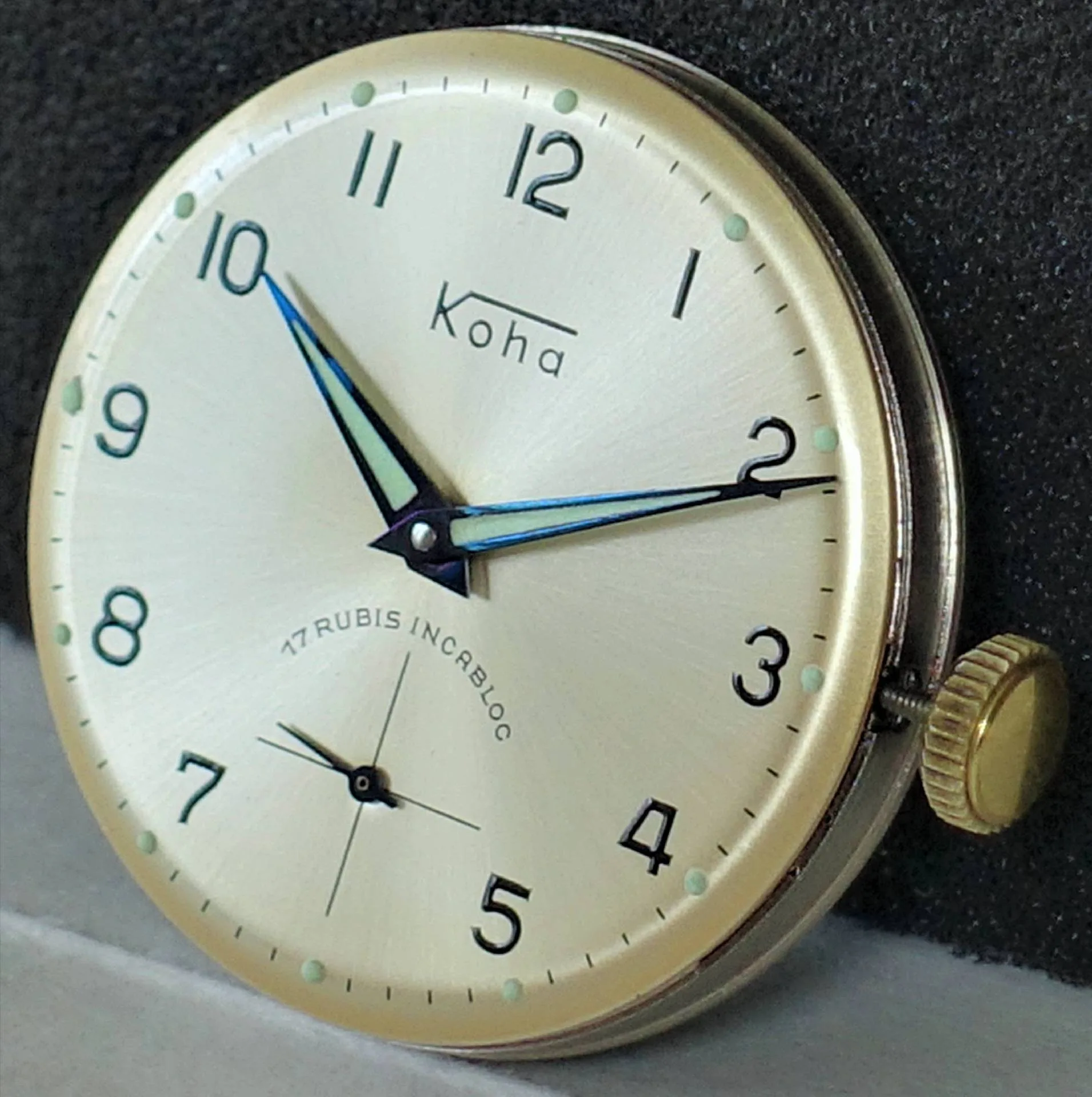 Koha Armbanduhr Zifferblatt Helles Zifferblatt einer Koha Armbanduhr mit schwarzen Zahlen und blauen Zeigern.