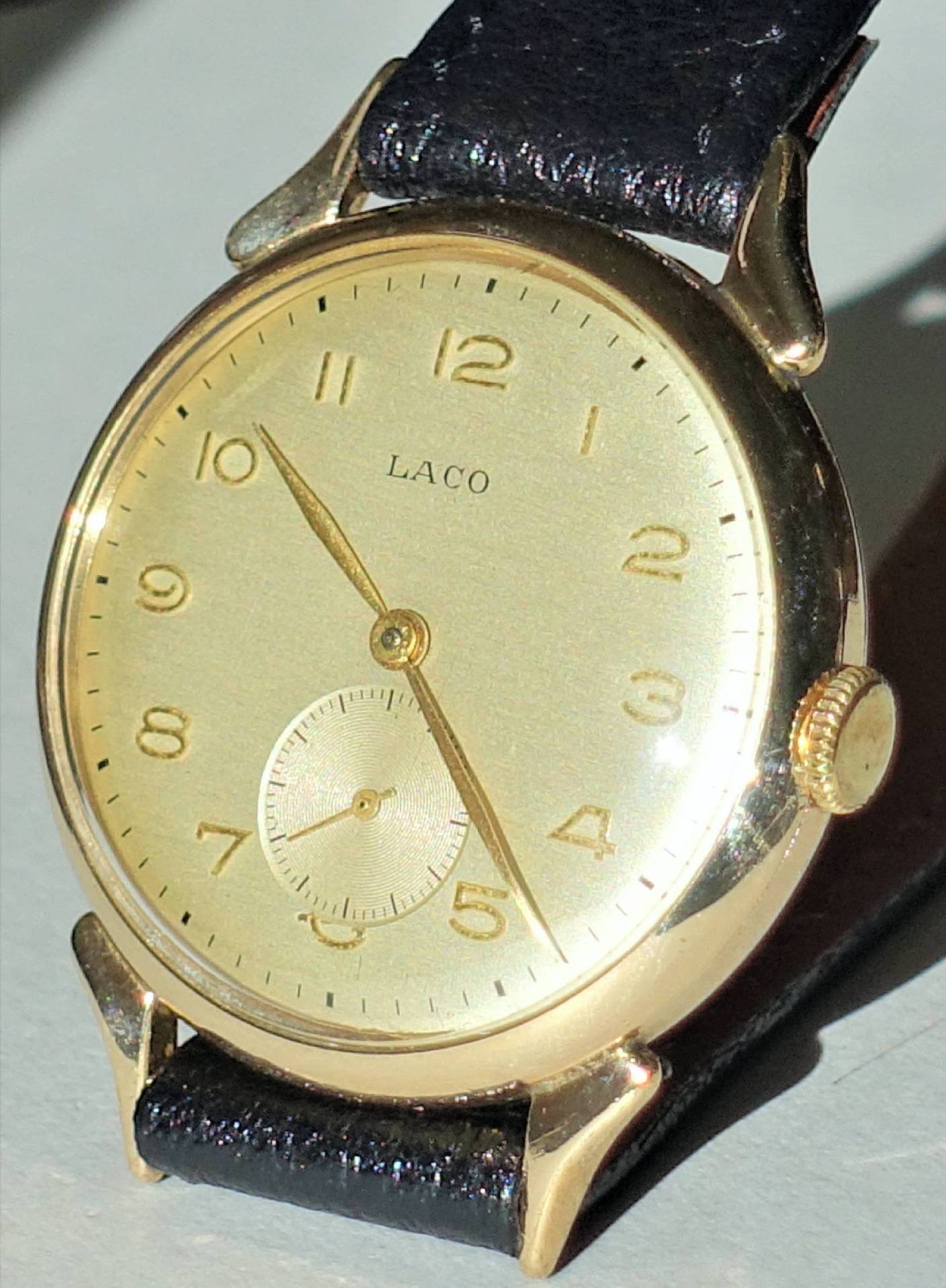 Goldfarbene Armbanduhr mit hellem Zifferblatt, schwarzen Ziffern und schwarzem Armband.