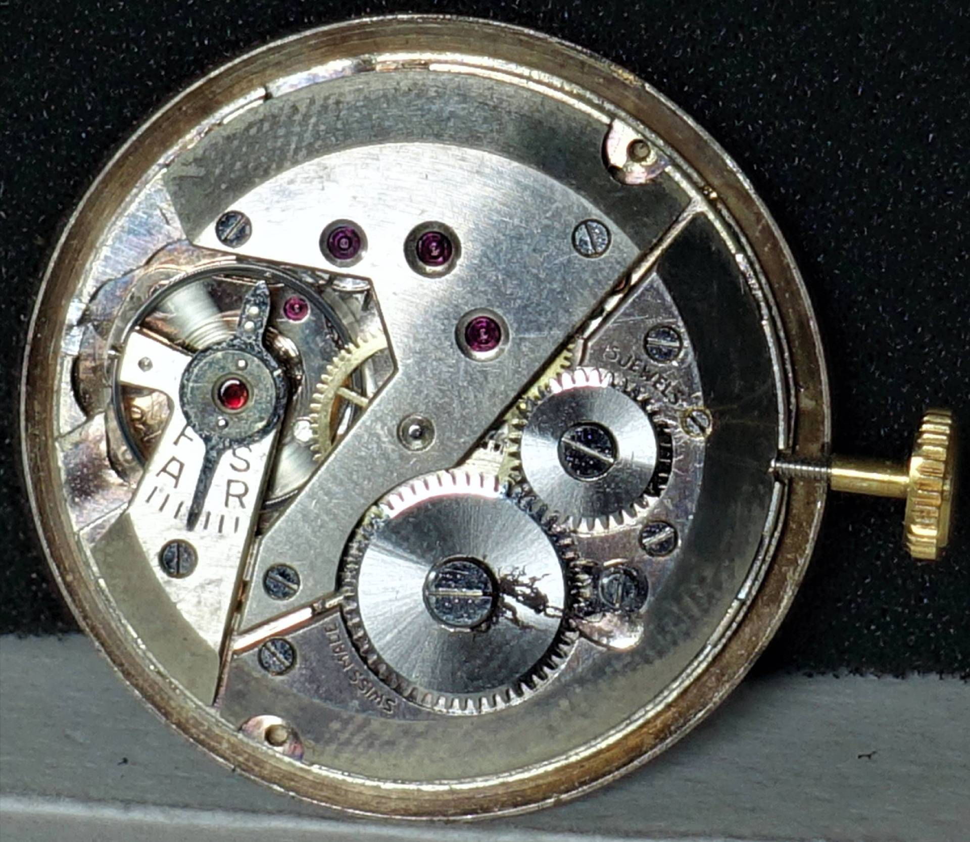 Silberfarbenes mechanisches Uhrwerk mit sichtbaren Zahnrädern und goldfarbener Krone.