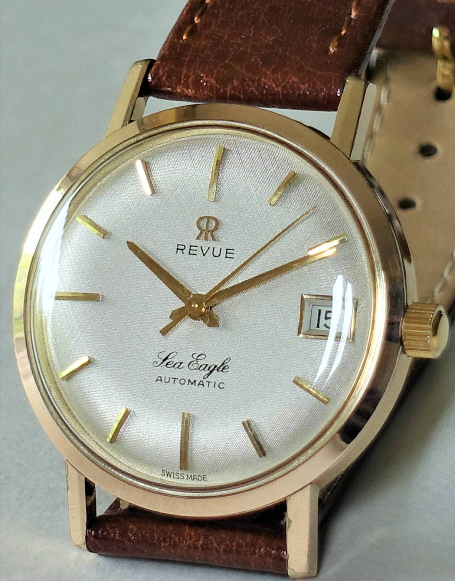 Goldfarbene Armbanduhr mit weißem Zifferblatt, braunem Lederarmband und Datumsanzeige.
