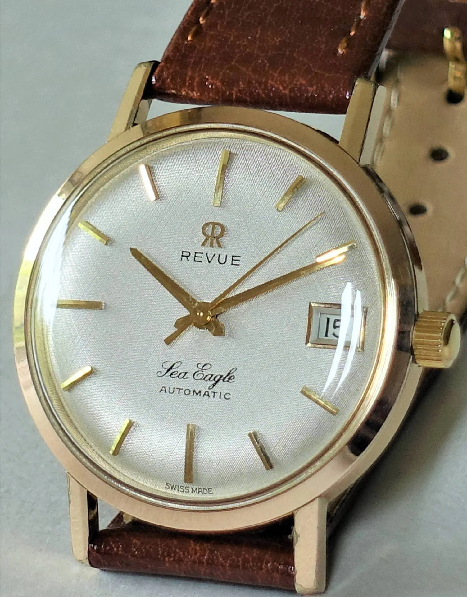 Goldfarbene Armbanduhr mit weißem Zifferblatt, braunem Lederarmband und Datumsanzeige.