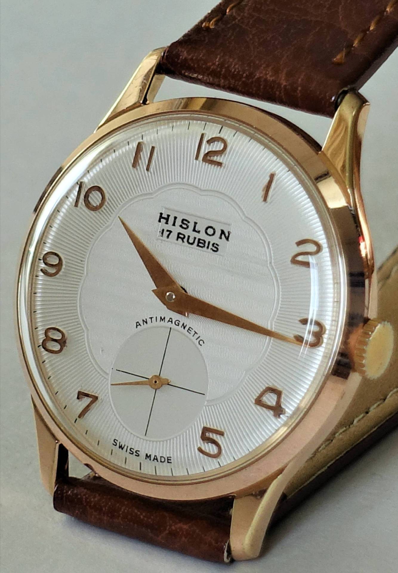 Eine klassische Armbanduhr mit weißem Zifferblatt, goldfarbenem Gehäuse und braunem Lederarmband, die Marke "HISLON" und "17 RUBIS" sind auf dem Zifferblatt zu sehen.