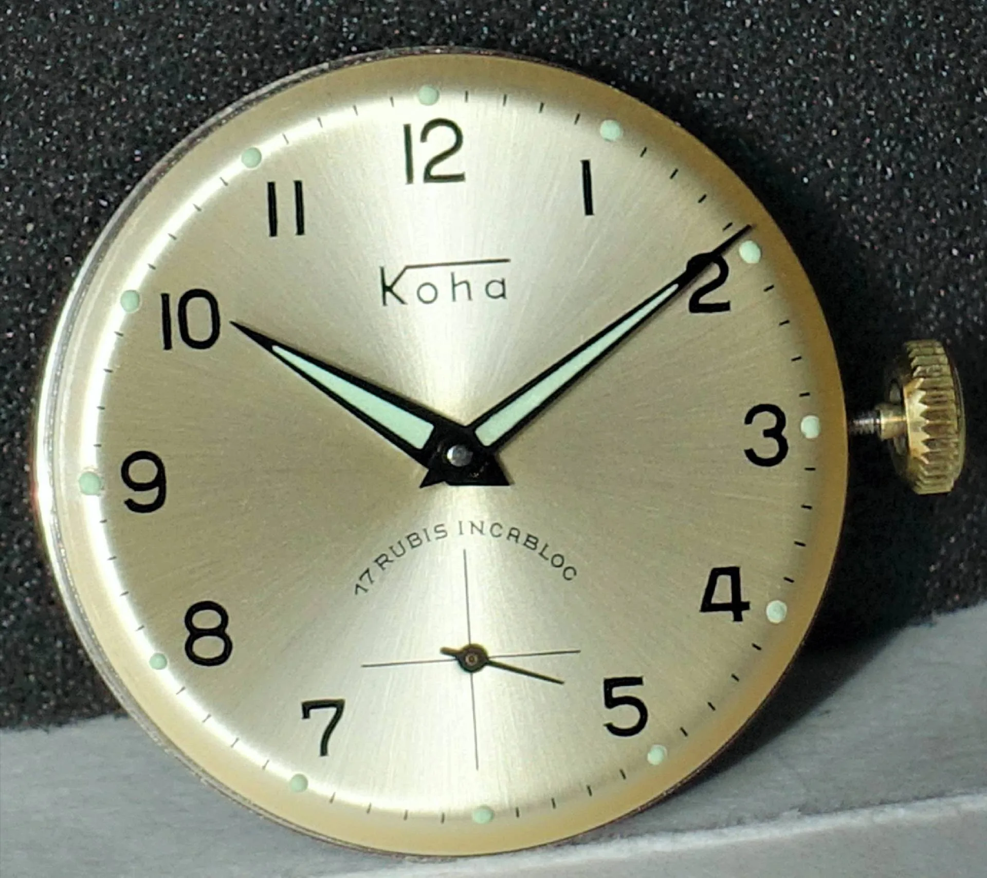Rundes goldfarbenes Uhrwerk mit hellem Zifferblatt, schwarzen Zahlen und Leuchtzeigern.