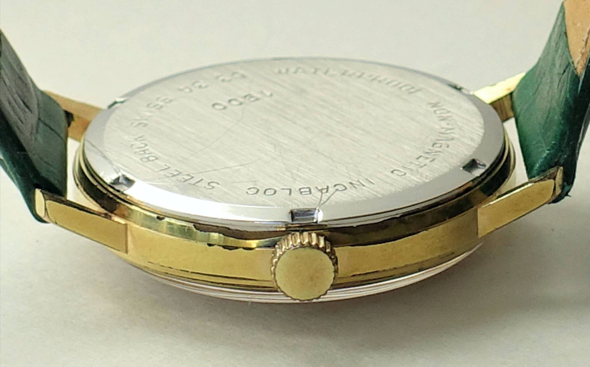 Rückseite eines goldfarbenen Uhrgehäuses mit silberfarbenem Boden und grünen Armbändern.