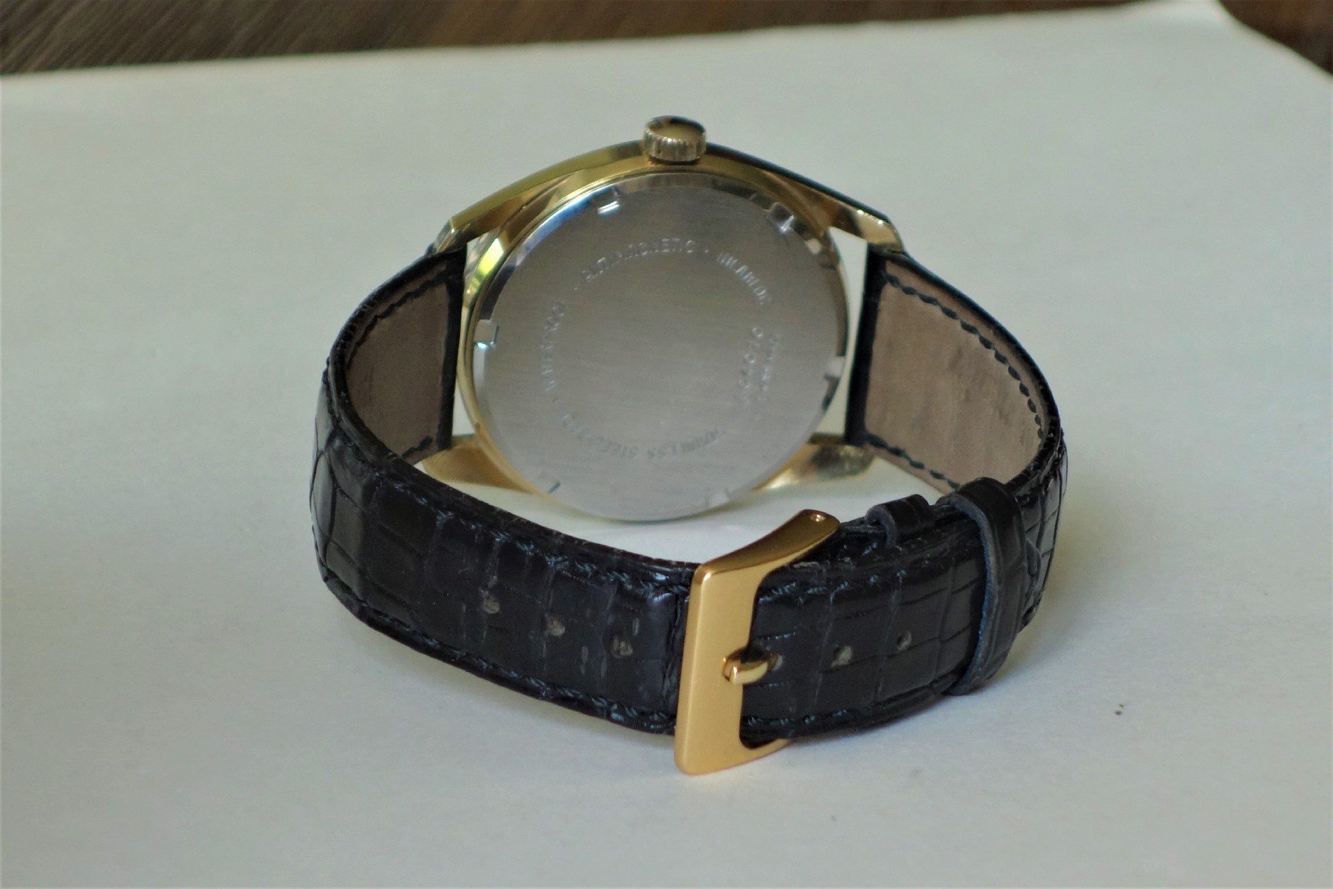 Analoge Armbanduhr mit schwarzem Lederarmband, goldfarbenem Gehäuse und silberner Rückseite.