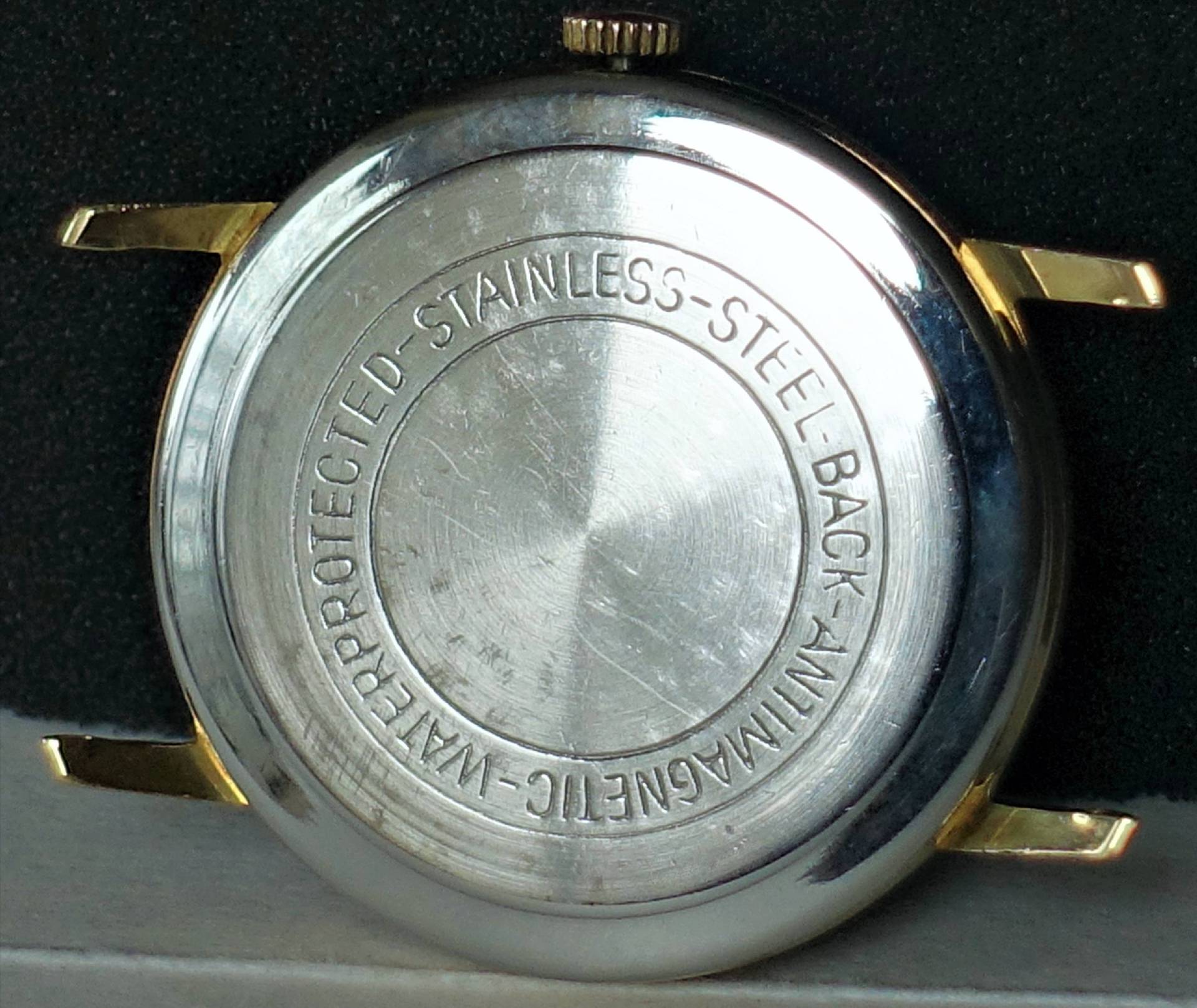 Silberfarbenes Uhrgehäuse mit gravierter Rückseite und goldfarbenen Bandanstößen.
