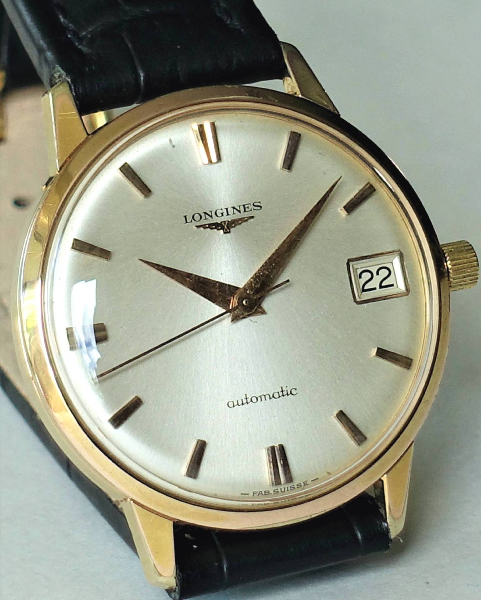 Goldfarbene Armbanduhr mit hellem Zifferblatt, schwarzem Armband und Datumsanzeige.