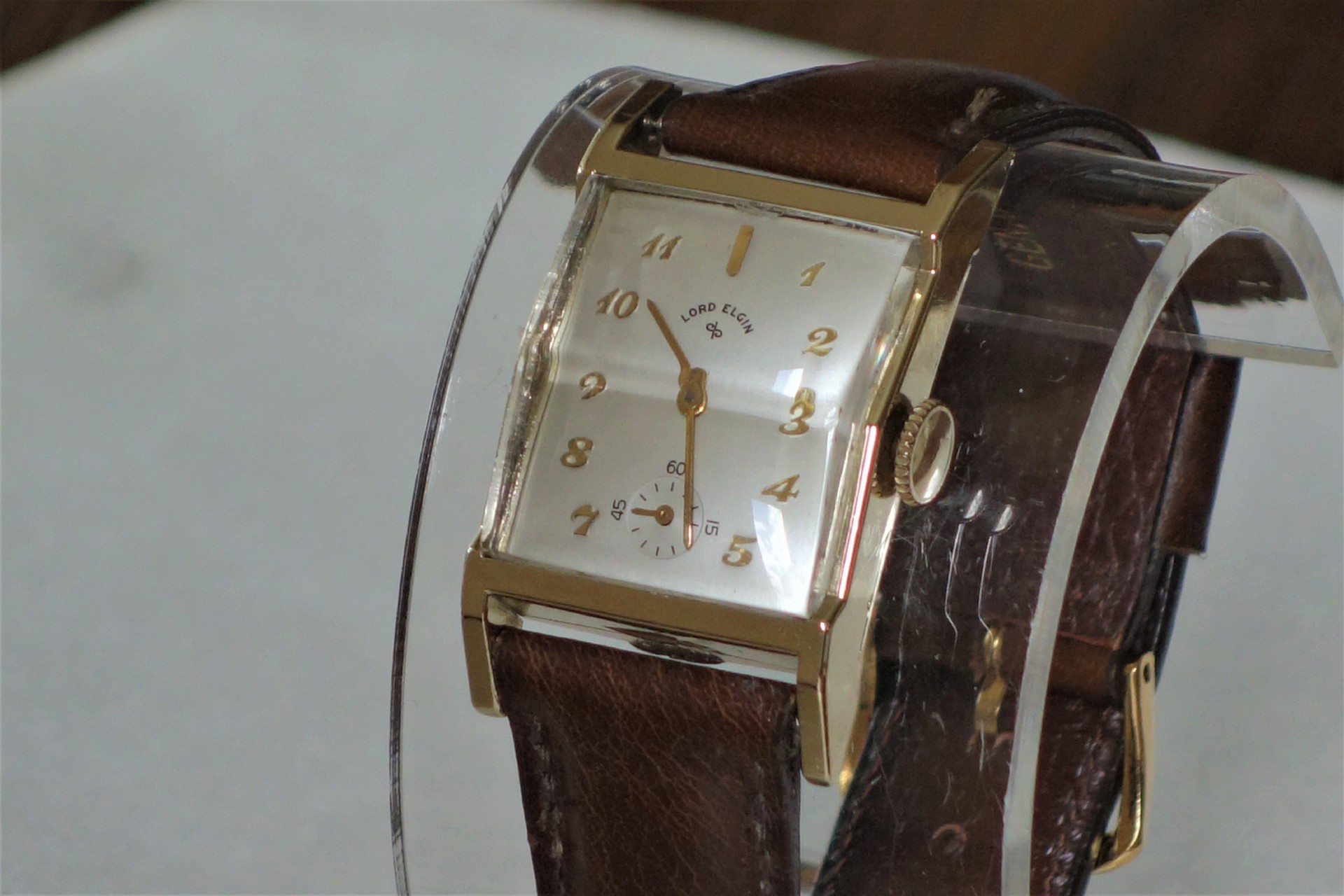 Rechteckige Armbanduhr mit braunem Armband und goldfarbenem Gehäuse.