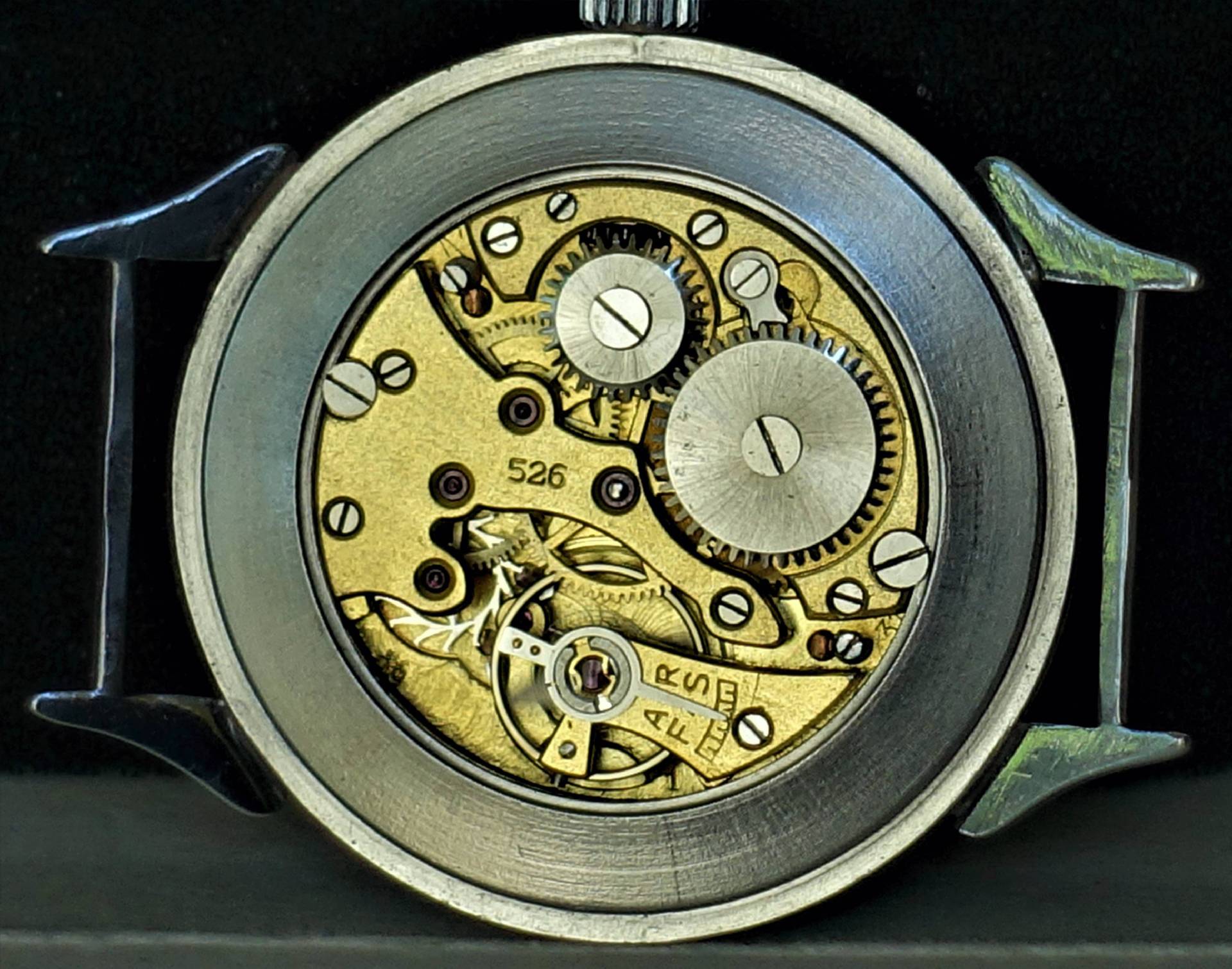 Mechanisches Uhrwerk in goldfarbener Ausführung mit sichtbaren Zahnrädern und Schrauben in einem silberfarbenen Gehäuse.
