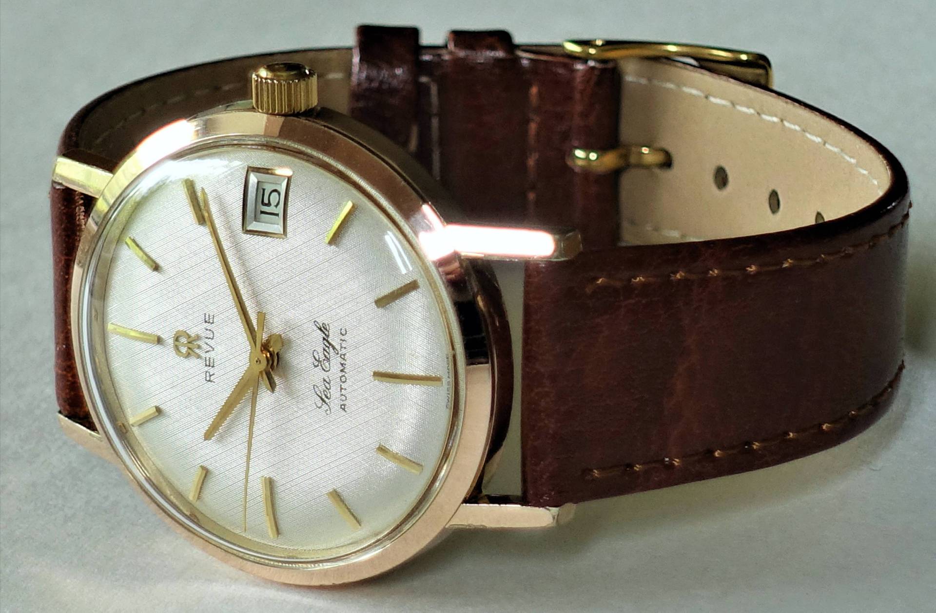 Goldfarbene Armbanduhr mit weißem Zifferblatt und braunem Lederarmband.