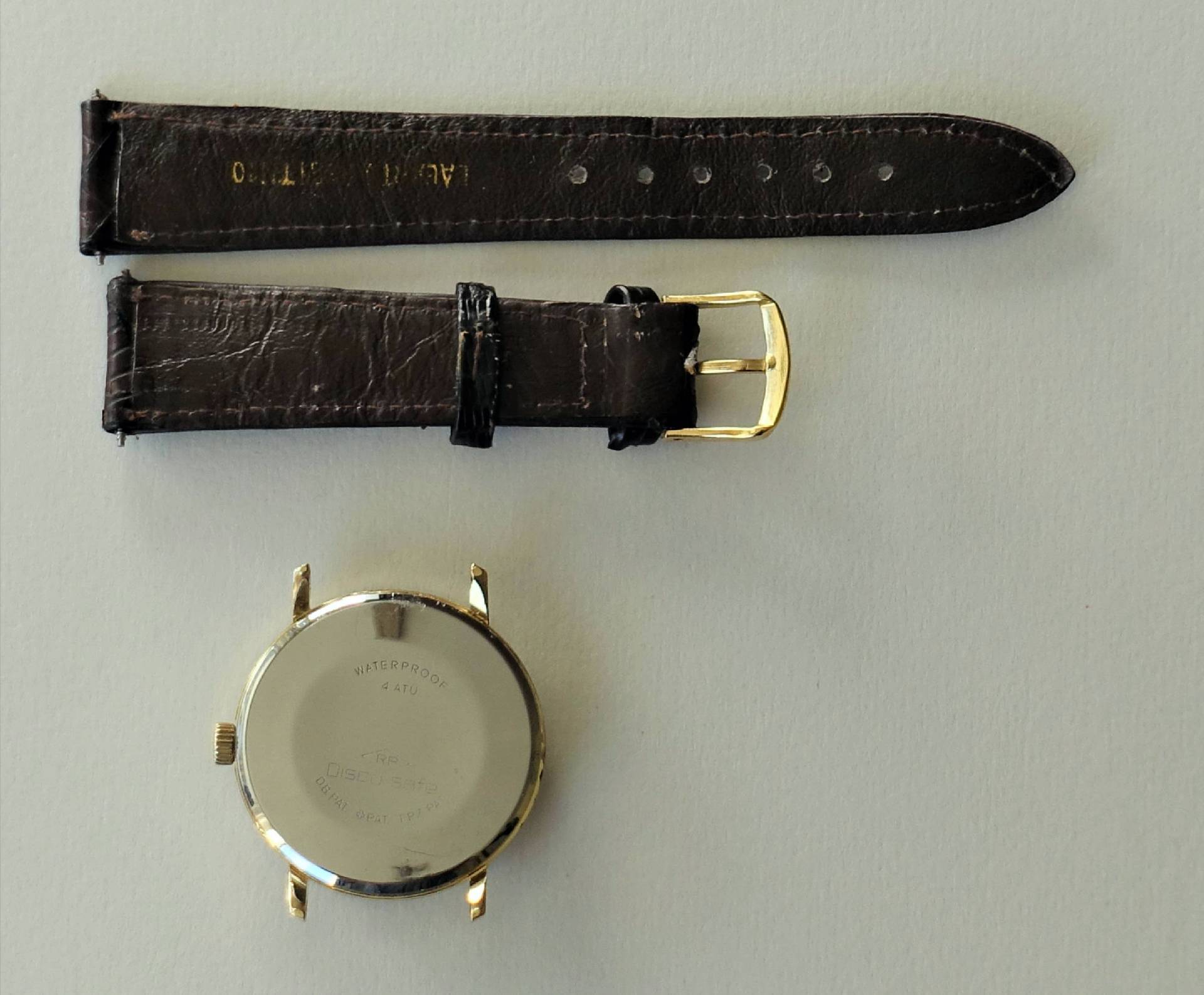 Zerlegtes goldfarbenes Uhrgehäuse mit braunem Lederarmband und goldfarbener Schließe.