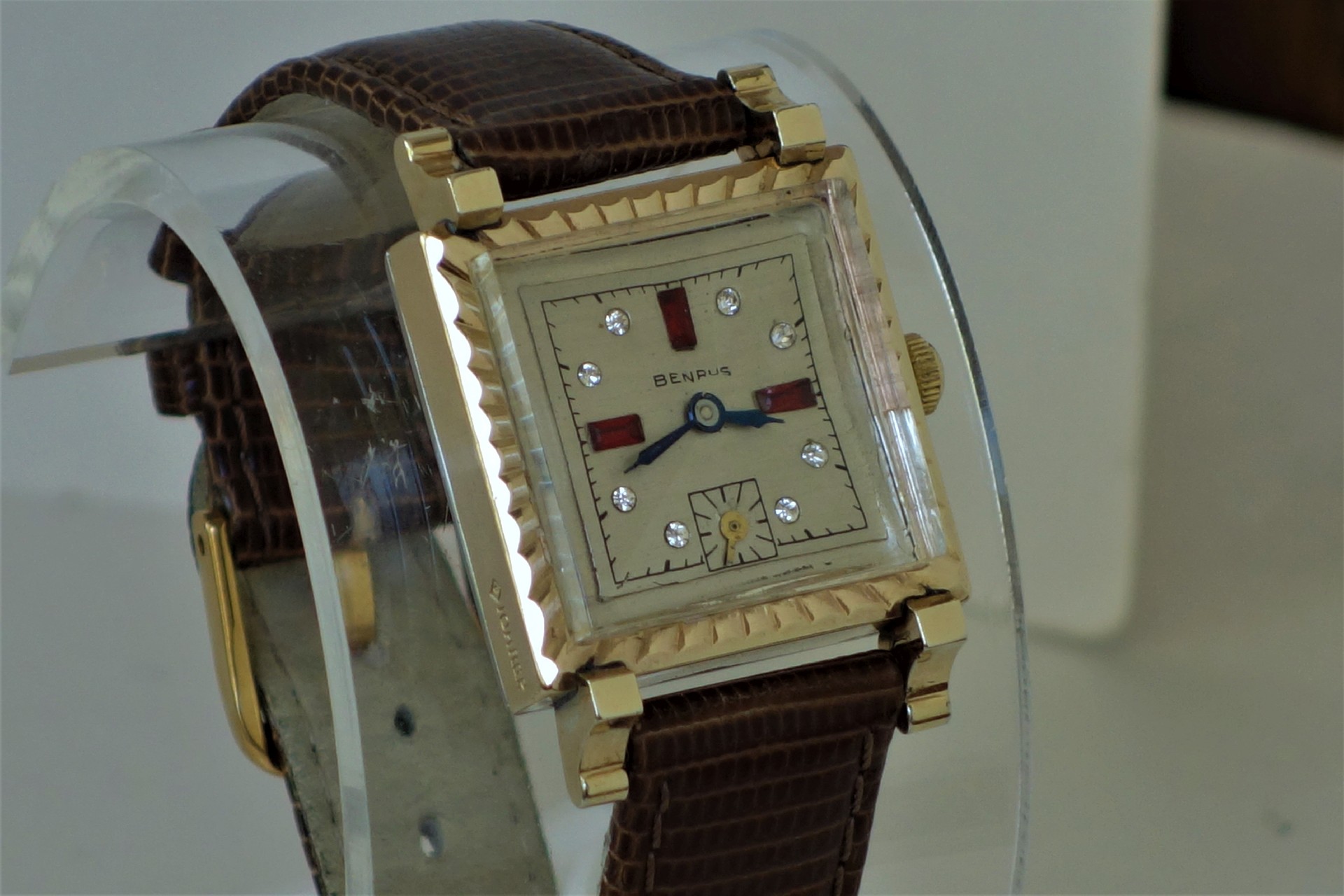 Rechteckige Armbanduhr mit goldfarbenem Gehäuse, hellem Zifferblatt, braunem Lederarmband und diamantähnlichen Indizes.
